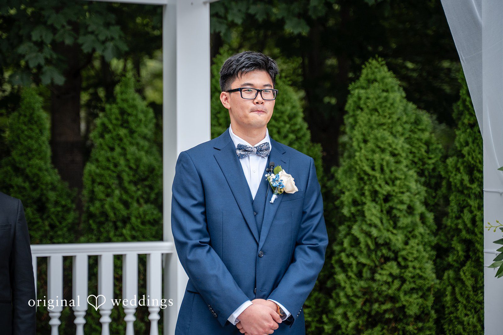 Groom in a blue suit standing alone in front of green hedges.