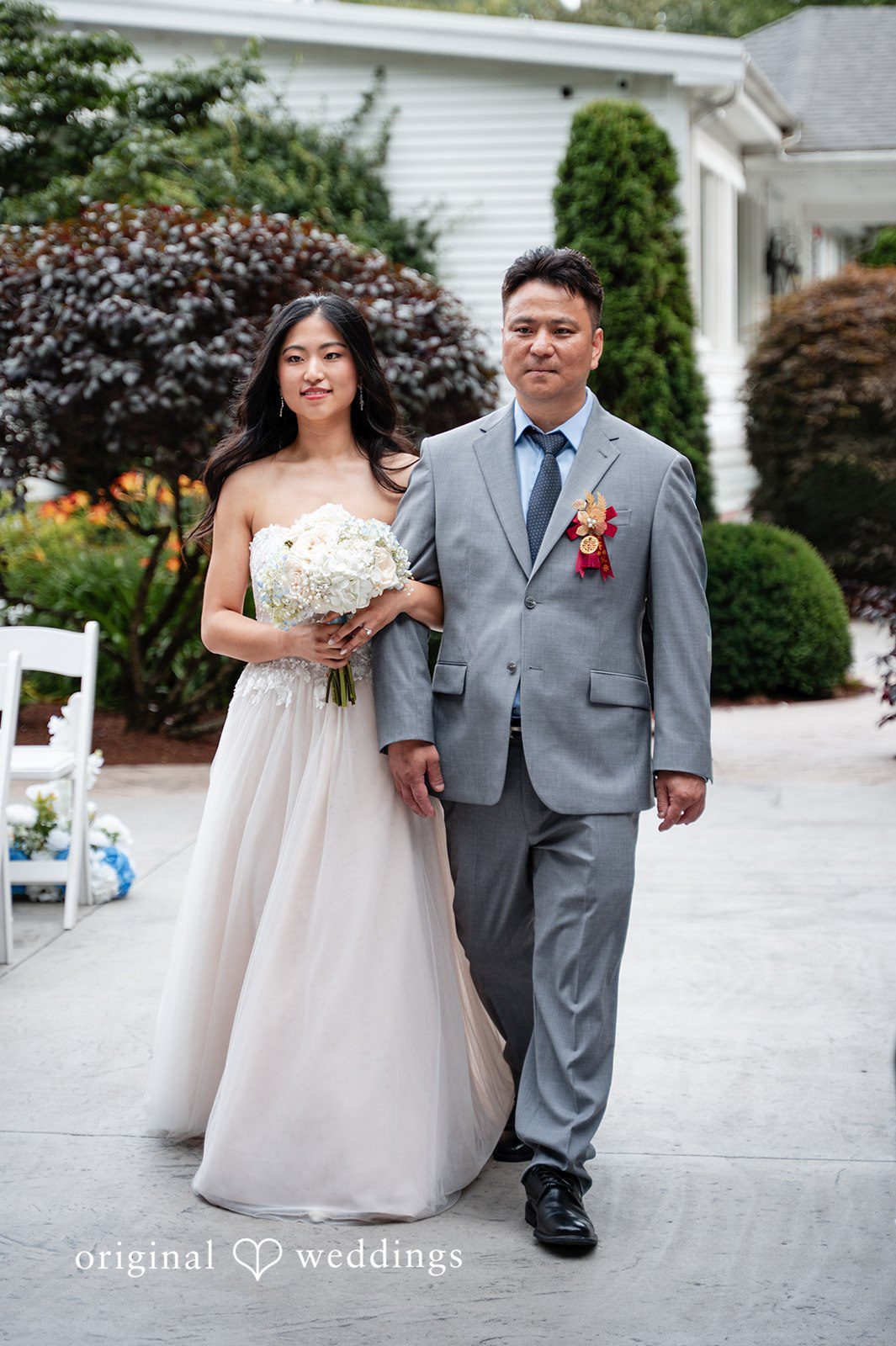 Bride in a strapless gown walking arm-in-arm with a man in a grey suit during a wedding procession.