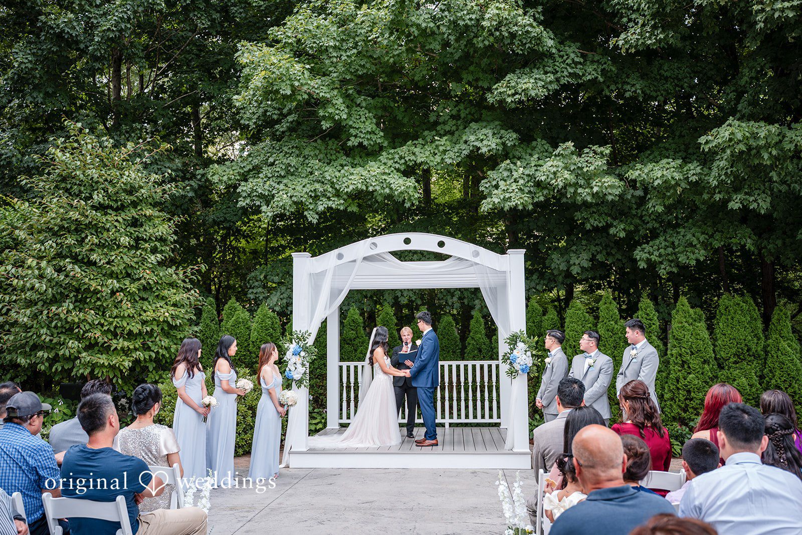 White wedding ceremony arch on a green lawn surrounded by trees.