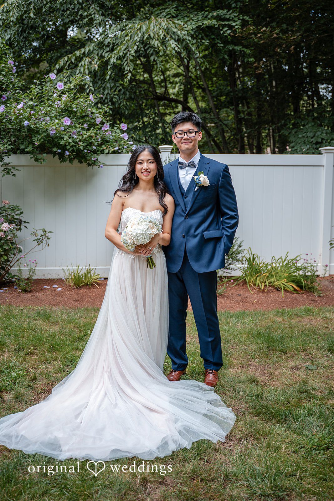 Bride in a flowing white gown posing on a lawn next to a groom in a dark suit.