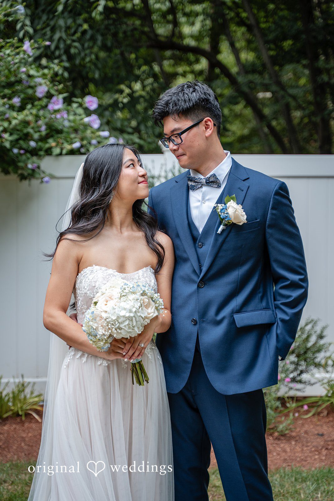 Bride and groom holding hands while posing together in front of greenery.