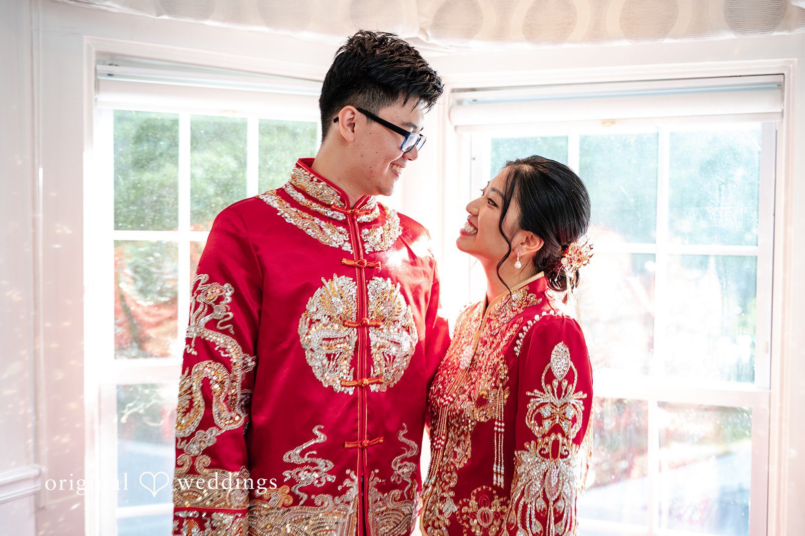 Bride and groom wearing traditional red Chinese wedding attire looking at each other near a window.