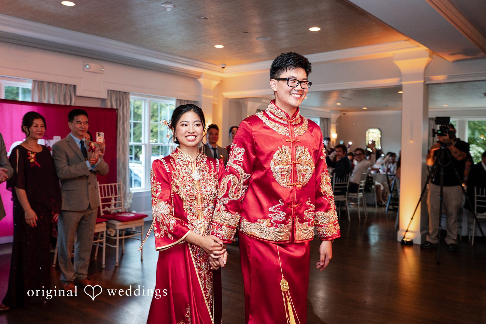 Bride and groom in red traditional garments walking through a banquet hall during a wedding event.