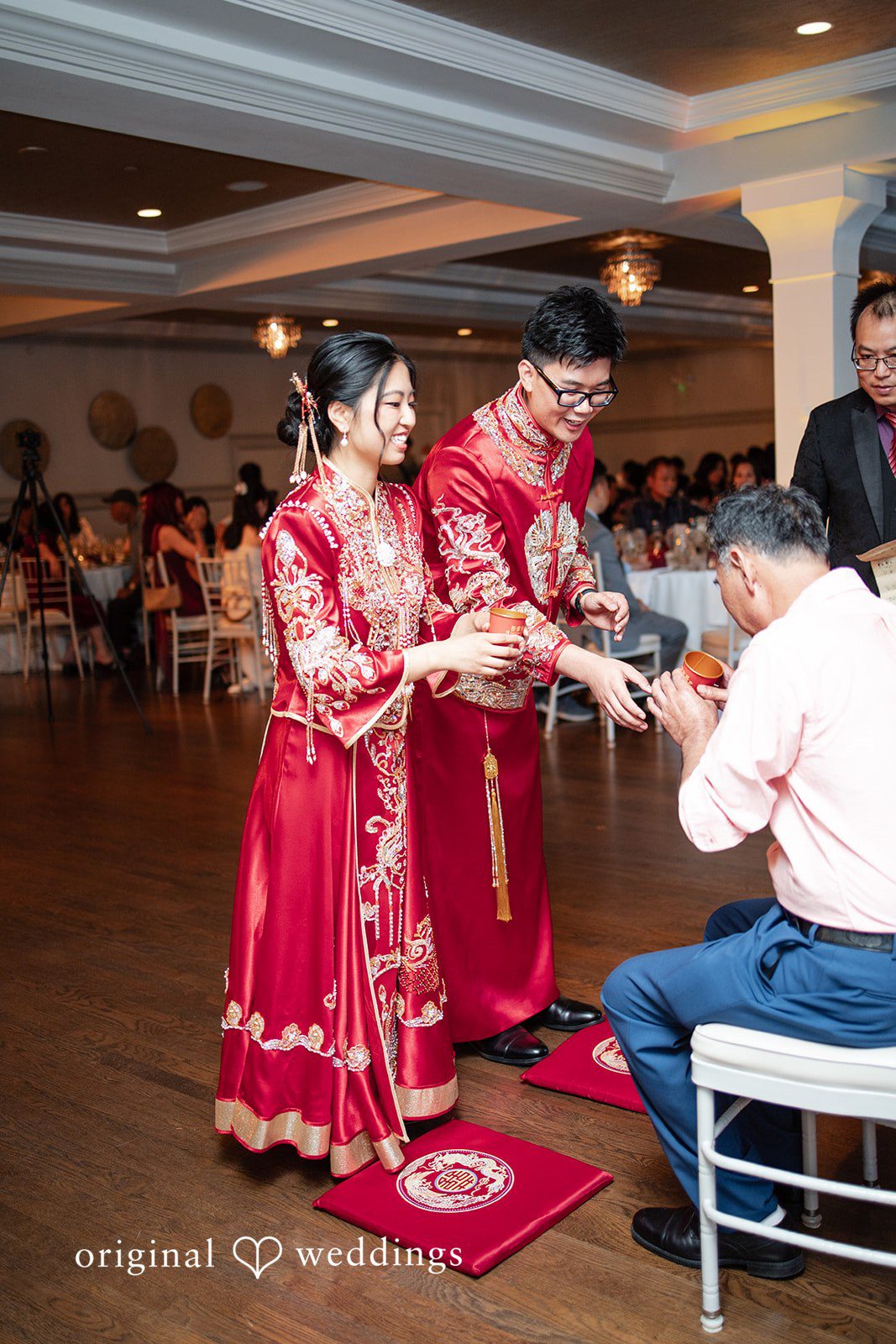 Bride and groom in red attire presenting a tea cup during a traditional Chinese tea ceremony.