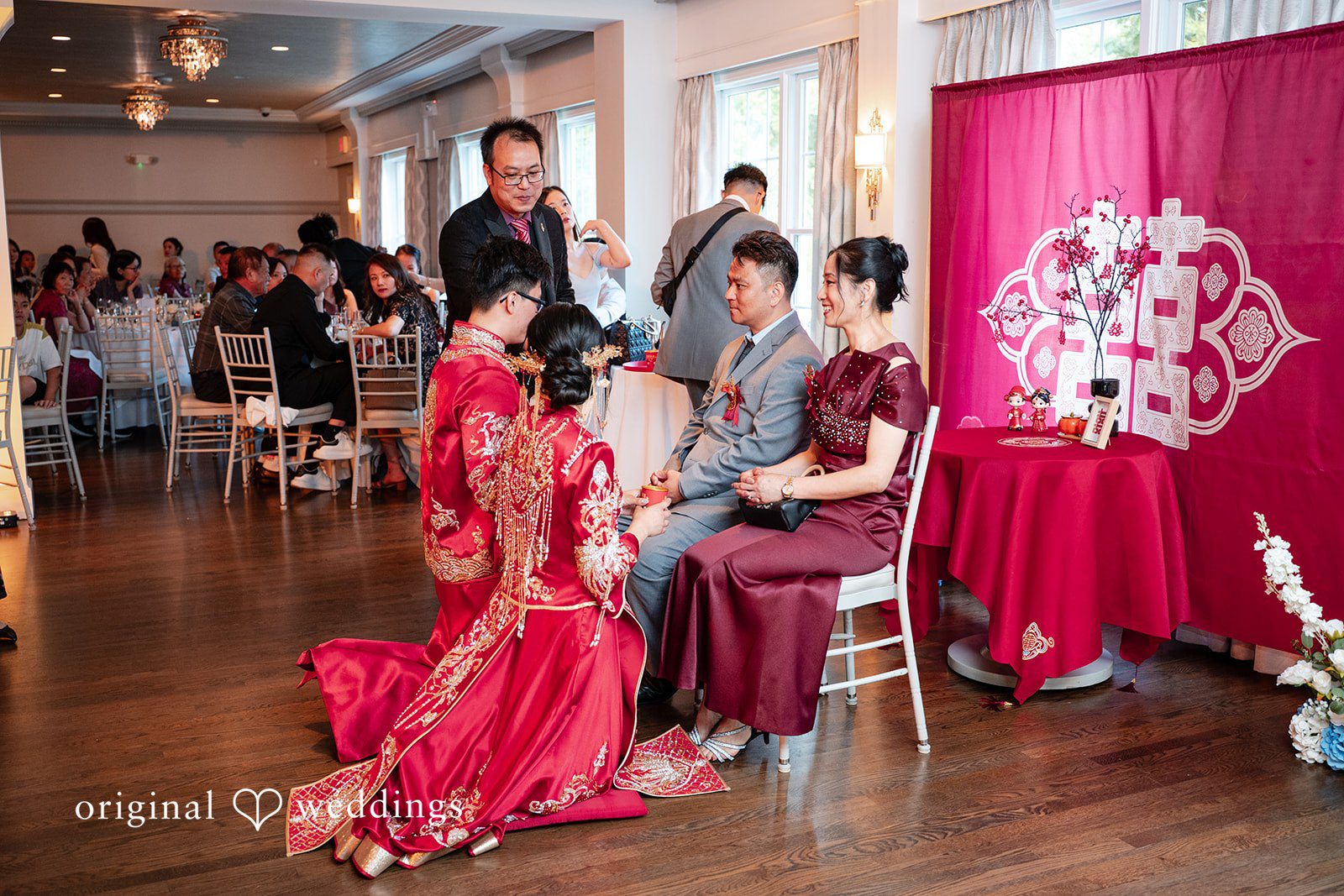 Bride and groom kneeling during a traditional tea ceremony in front of seated guests and a red backdrop.