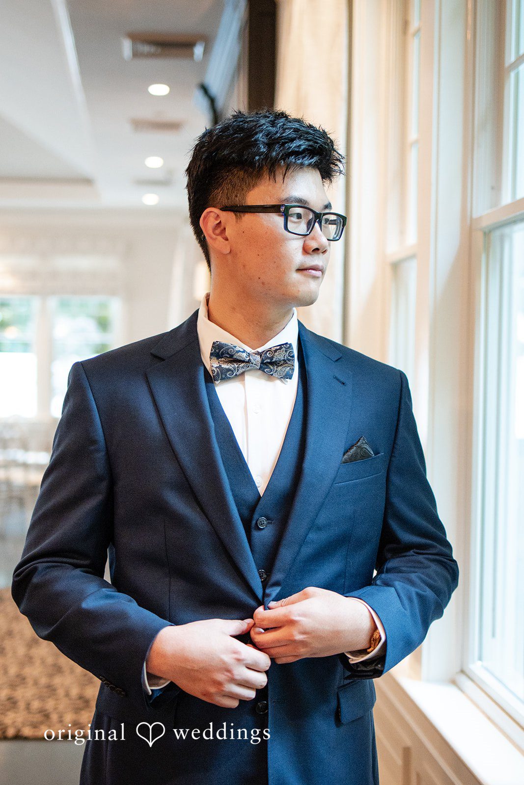 Groom in a dark suit and bowtie posing near a window.