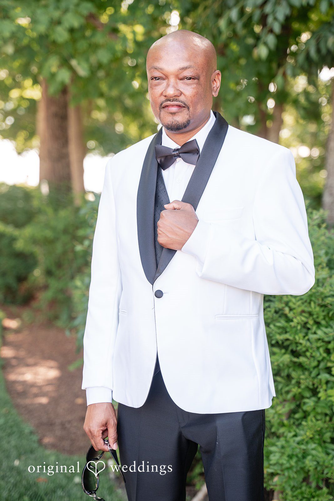 Melissa + Walter A groom wearing a white tuxedo jacket with a black bow tie, smiling broadly.