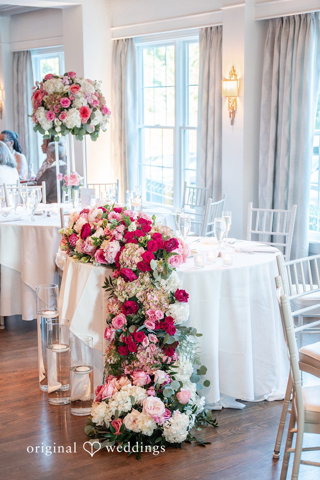 Melissa + Walter A wedding reception table decorated with a cascading floral arrangement of red, pink, and white flowers.