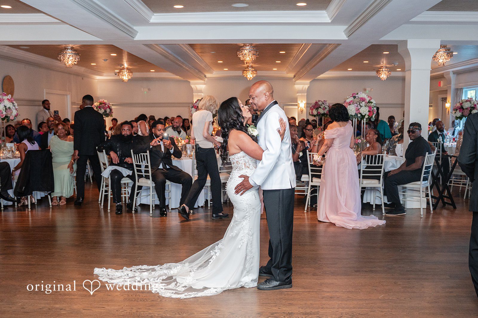 Melissa + Walter A bride and groom share their first dance in the center of a crowded wedding reception ballroom.