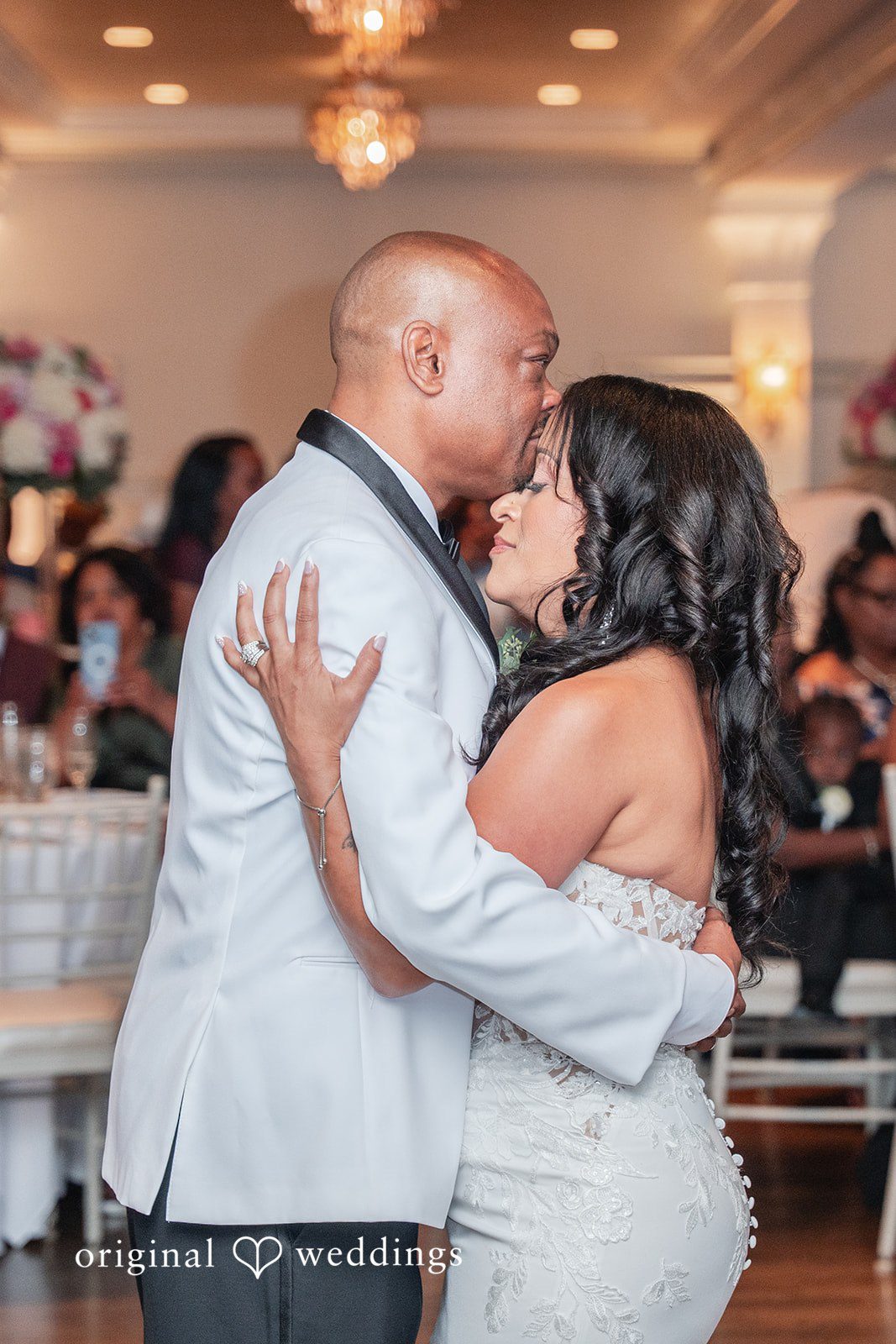 Melissa + Walter A groom embracing his bride during their first dance, with guests visible in the background.