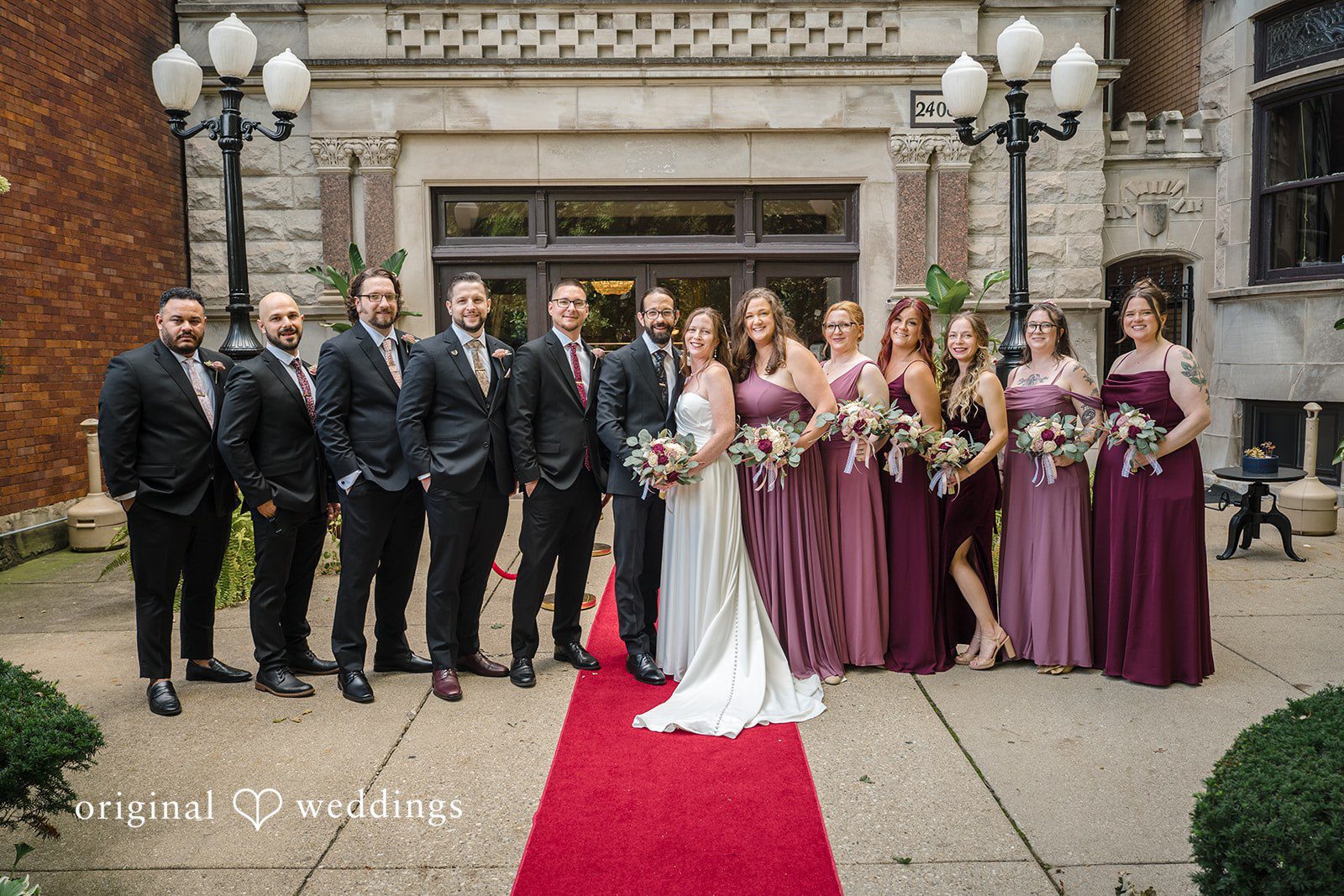 Kimberly + David Large bridal party posing together on a red carpet outdoors next to a brick building.