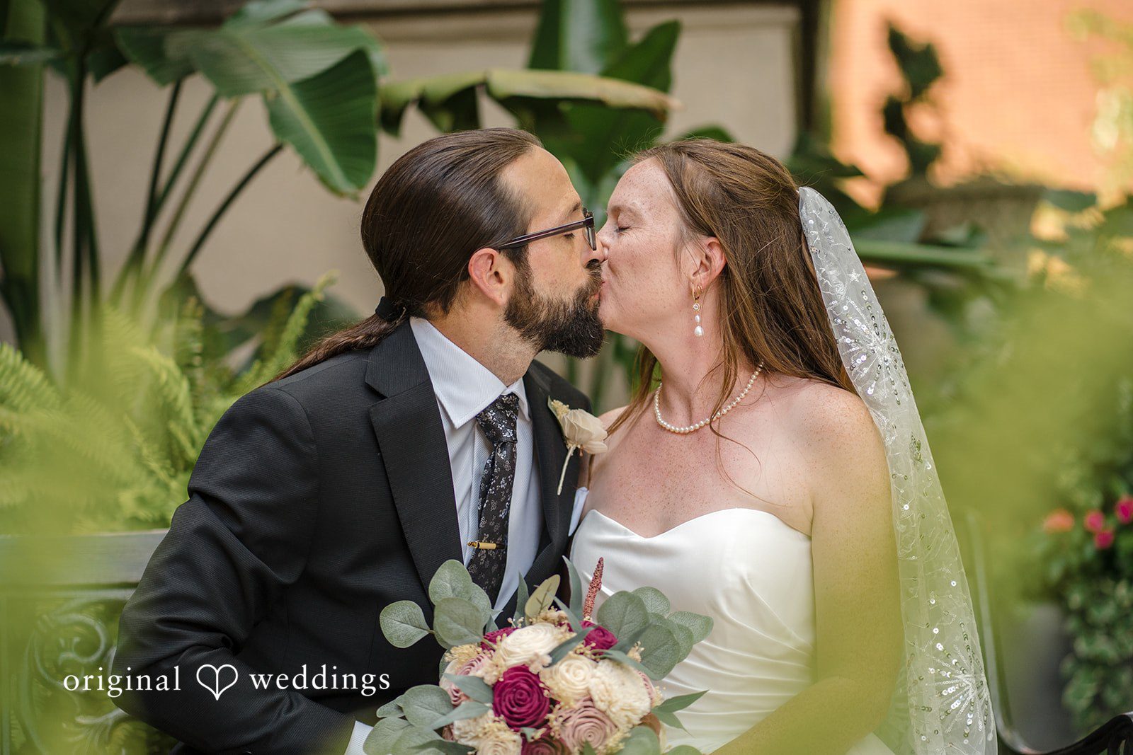 Kimberly + David Bride and groom kissing while seated on a bench surrounded by green tropical plants.