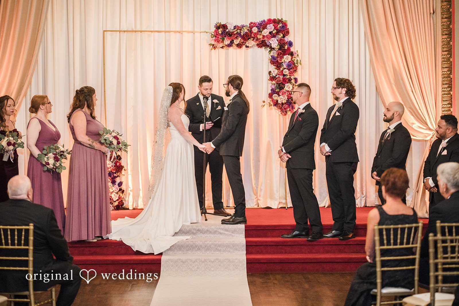 Kimberly + David A wedding ceremony in progress on a red stage with the bride and groom holding hands at the altar surrounded by their bridal party.