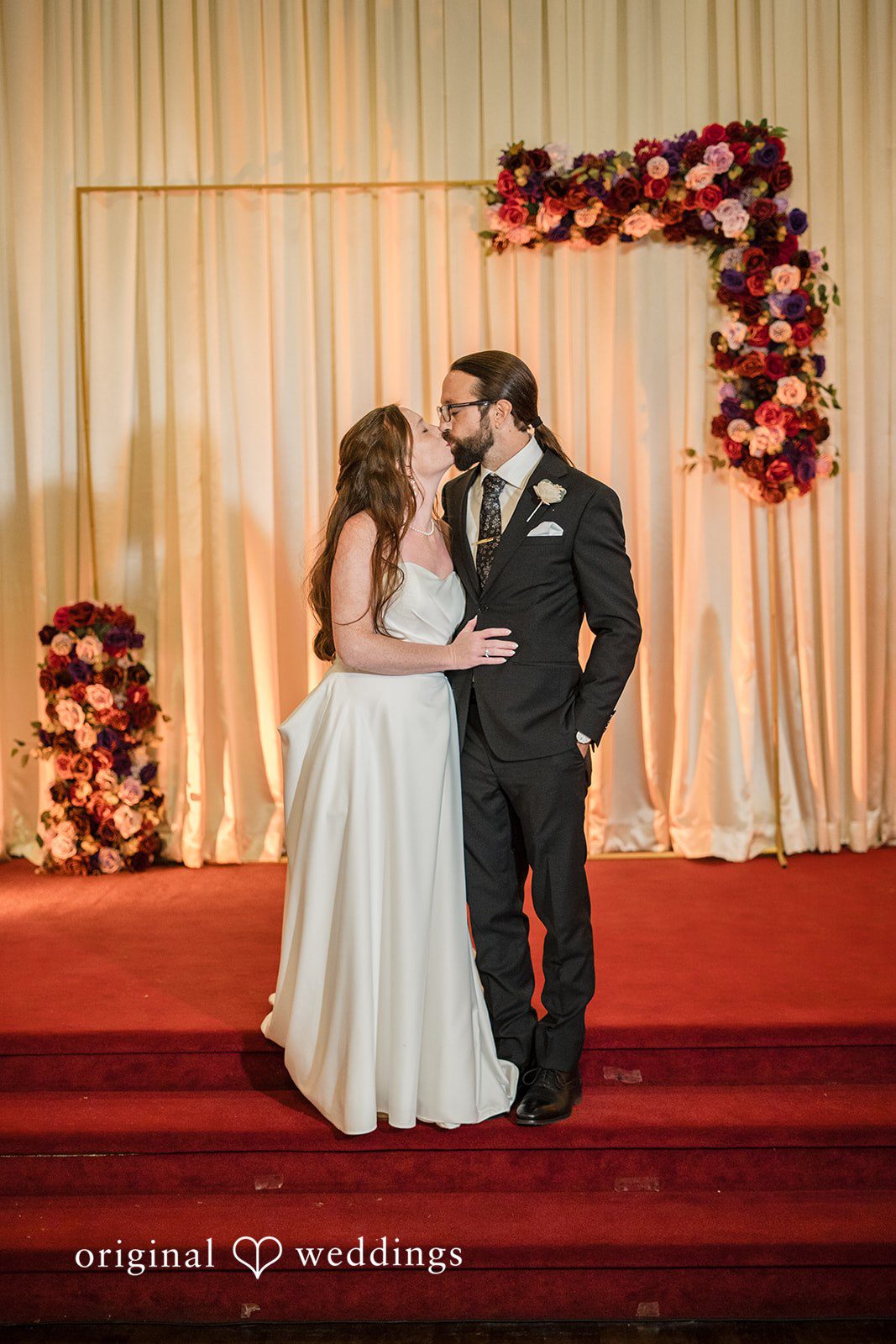 Kimberly + David Newlyweds kissing on a red carpet stage in front of a white curtain backdrop.
