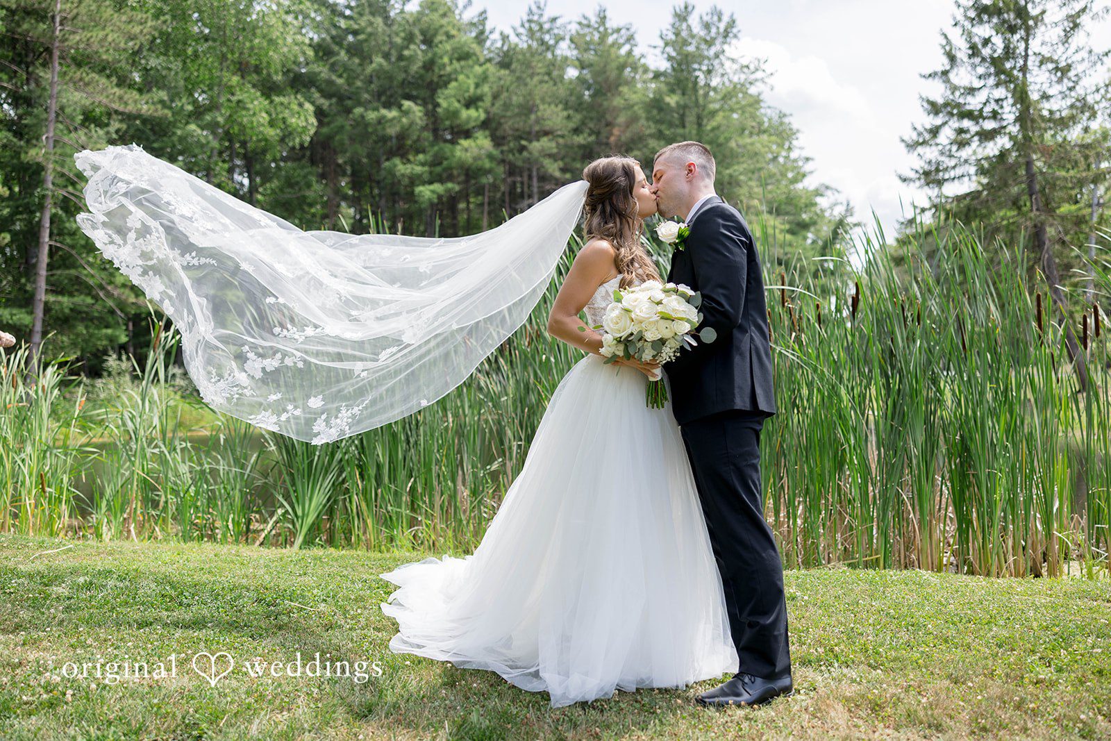 A bride with a long veil stands alongside a groom near a wooded area.