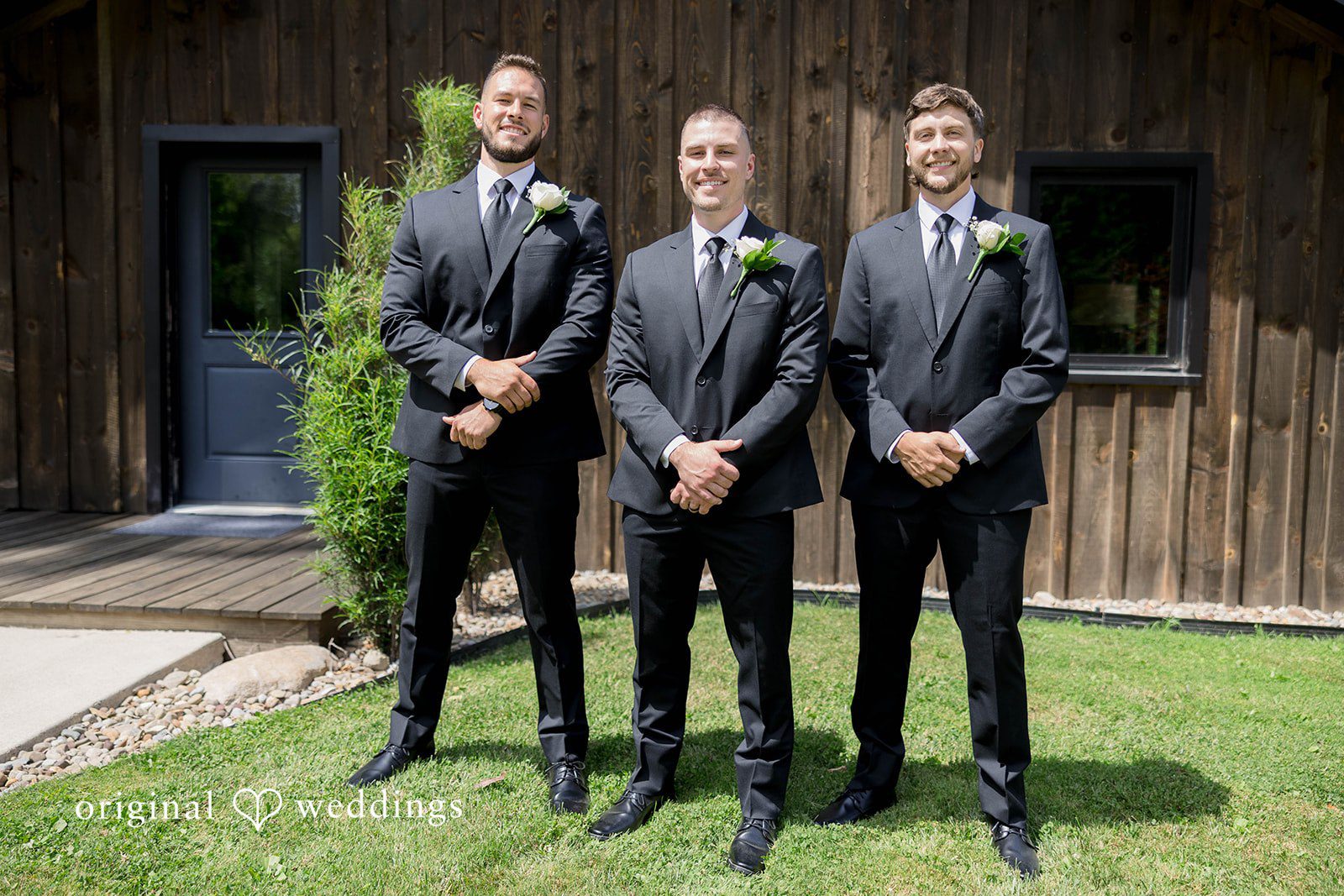 Three men in formal suits stand in a row against a wooden building.
