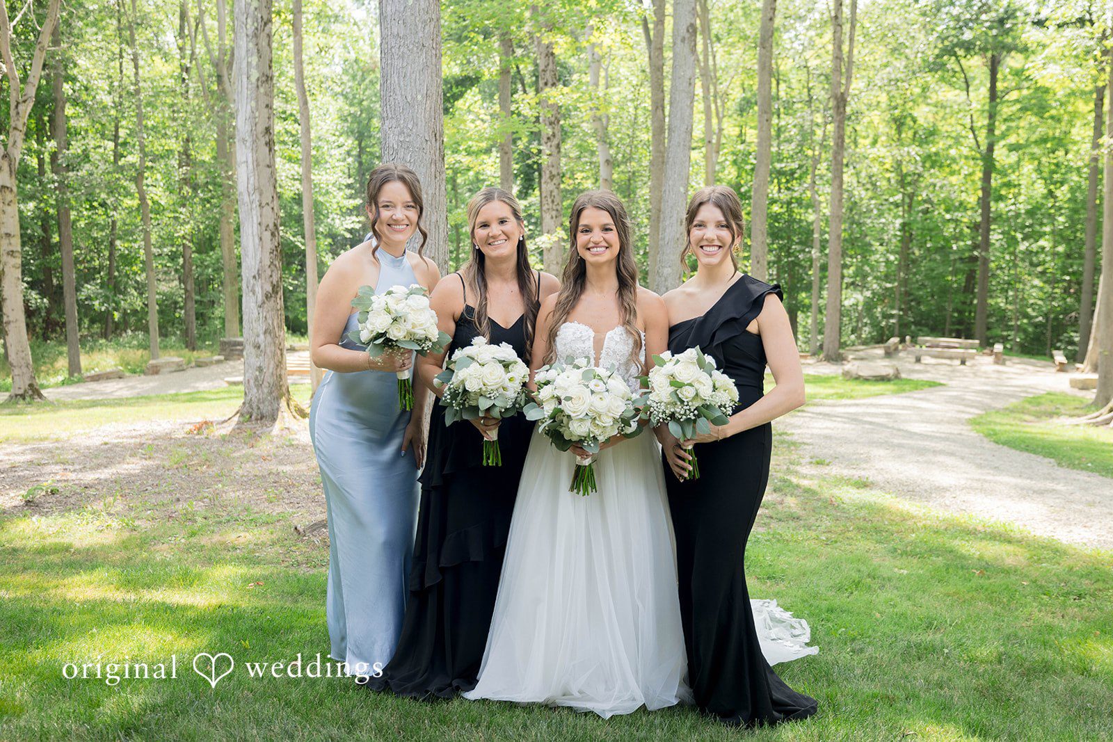 Bridesmaids in dark blue gowns stand with a bride along a tree-lined path.