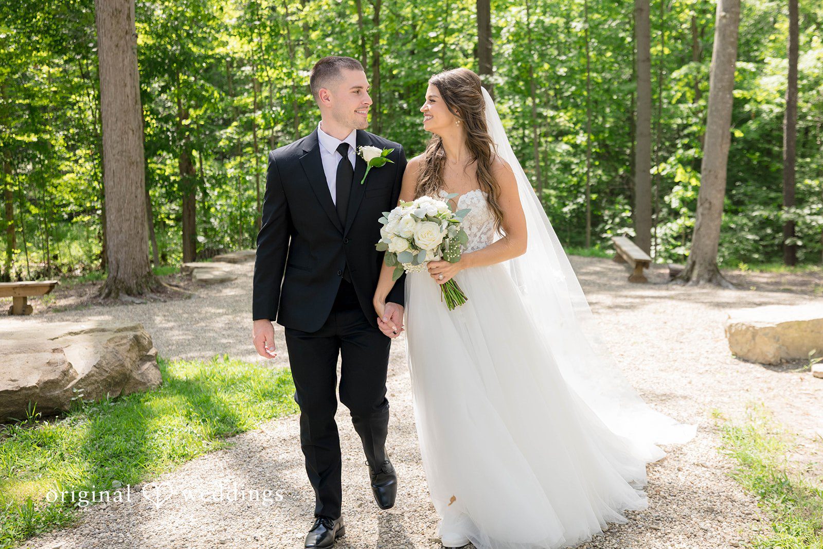 A bride and groom walk together along a path through the woods.