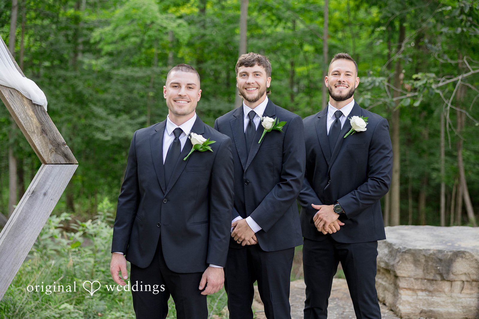 Groom and groomsmen in matching dark suits stand side-by-side in front of greenery.