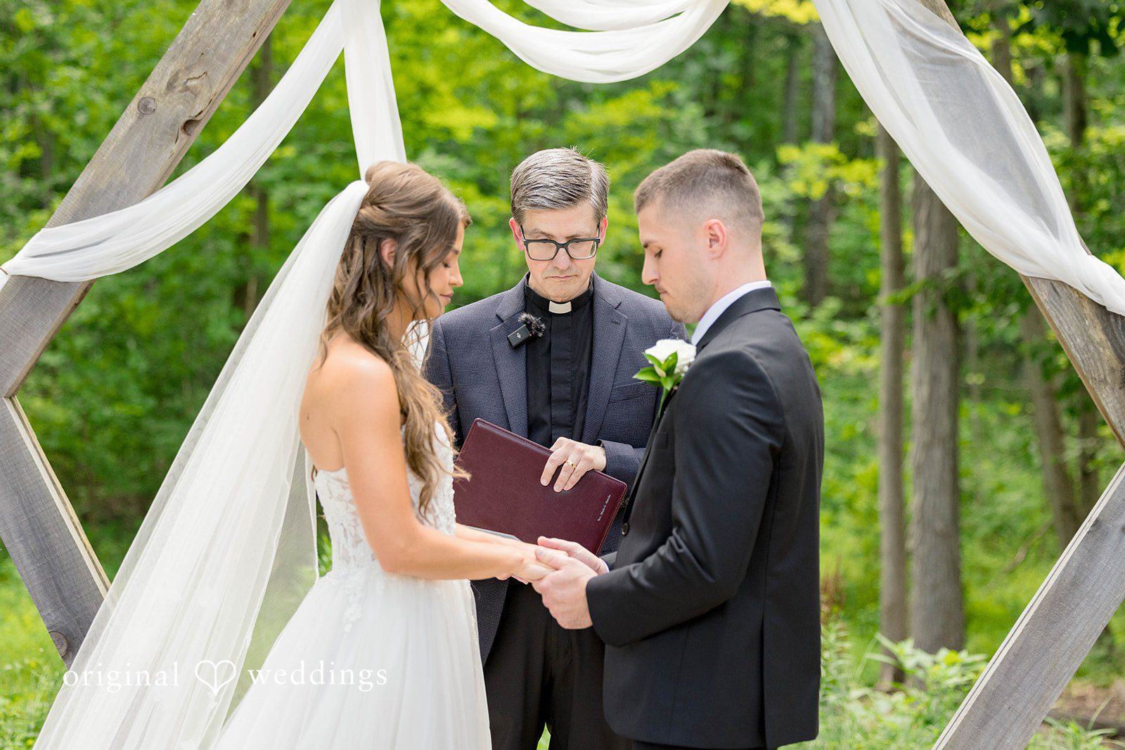 A wedding ceremony takes place beneath a draped white arbor frame.