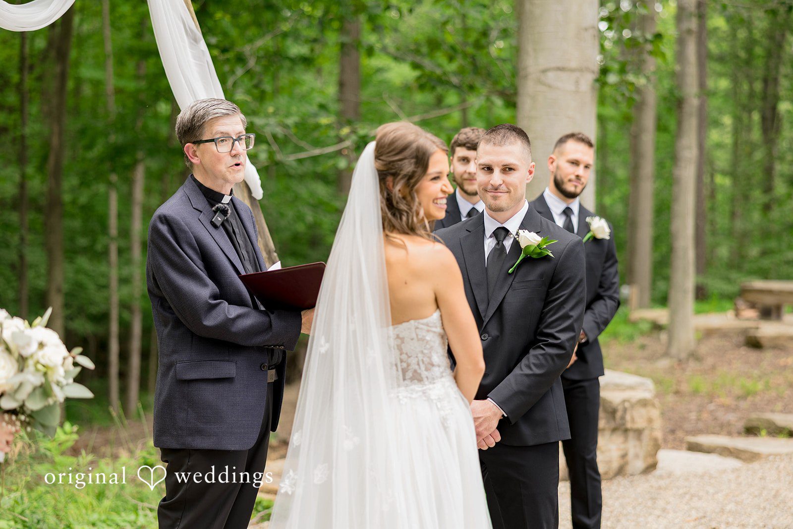An officiant in a dark jacket and glasses reads from a folder during a wedding ceremony while a bride with a long veil and a smiling groom in a black suit look on.