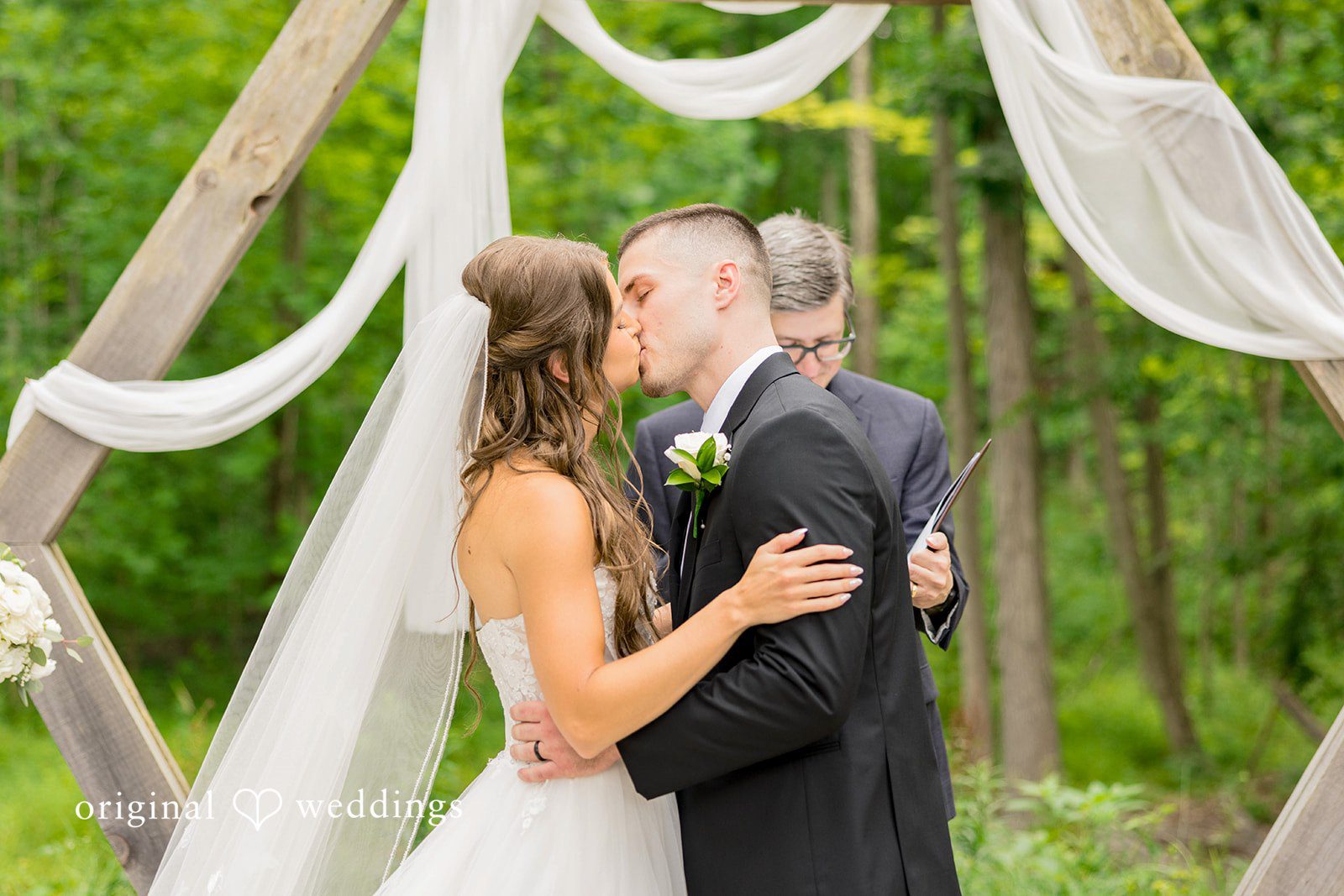 A bride with a long veil and a groom in a black suit share a first kiss beneath a wooden geometric arbor draped with flowing white fabric.