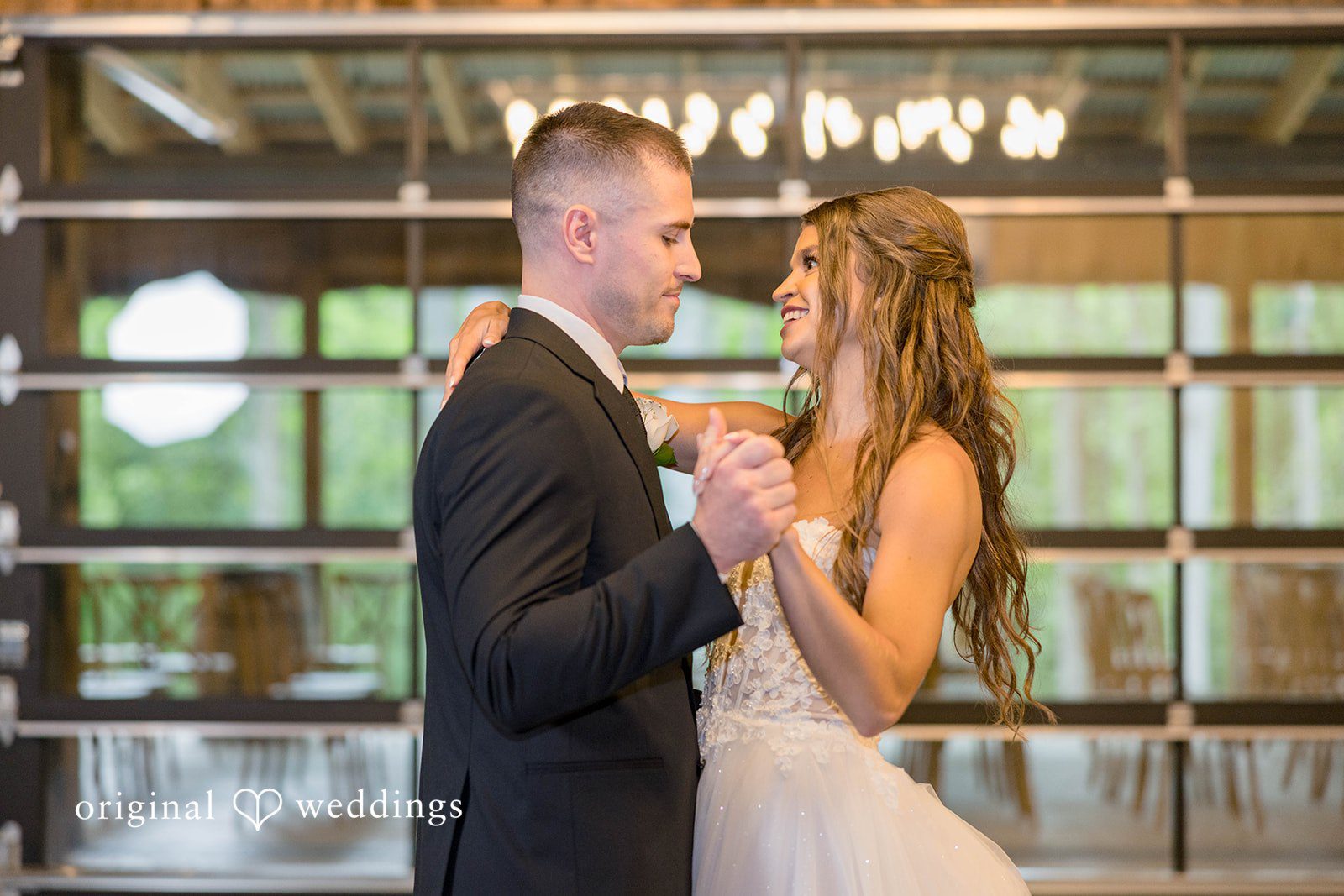 A bride and groom face each other while slow dancing in a well-lit reception space.