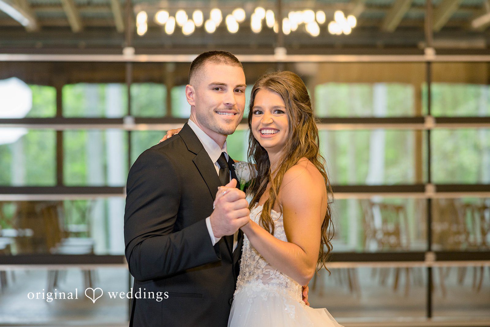 A bride and groom smile at the camera while holding hands on the reception dance floor.
