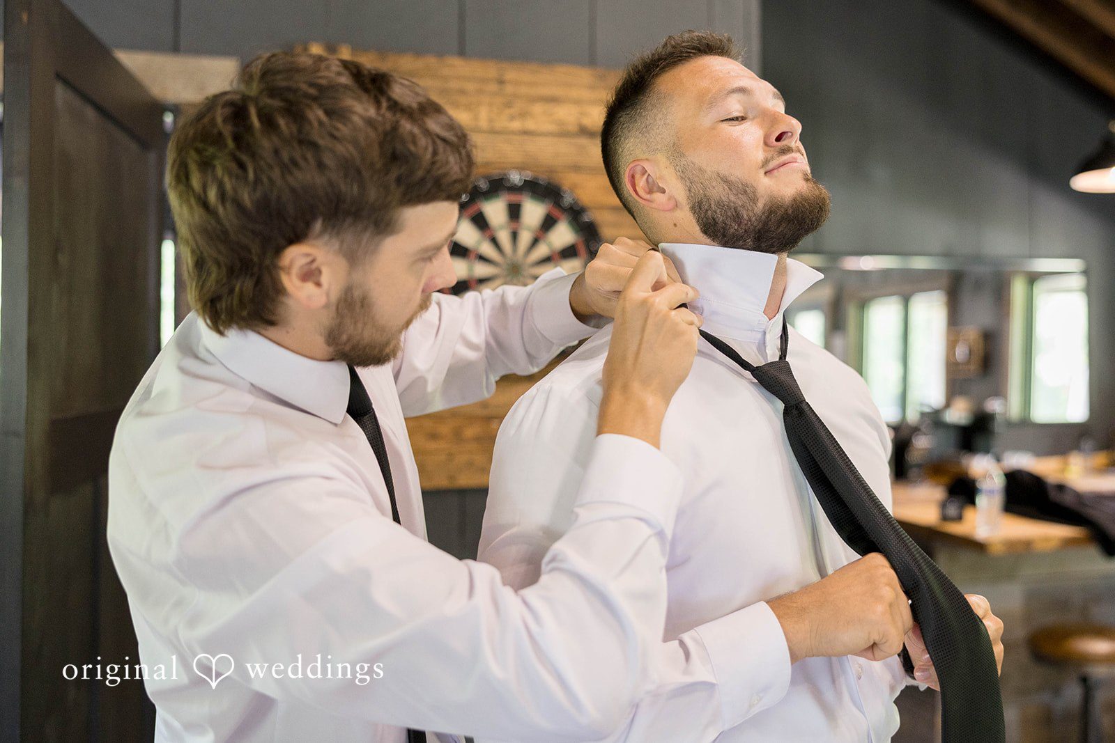 A man assists a groom with his vest and tie while getting ready.