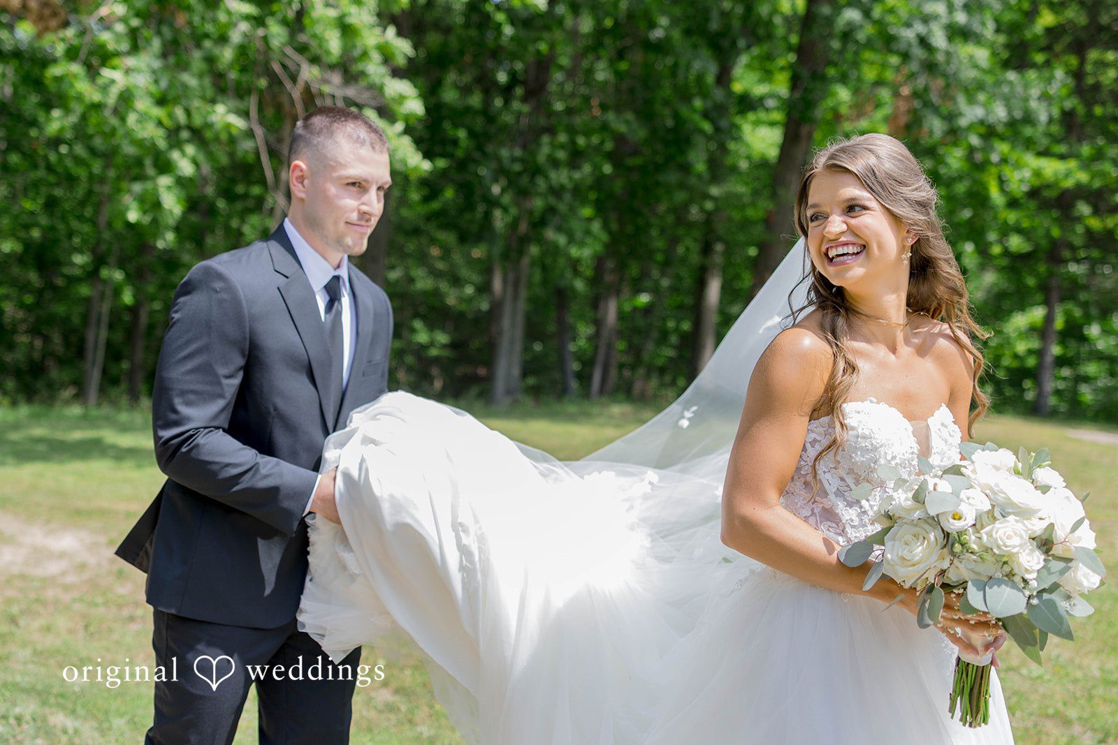 A bride in a long flowing veil stands with a groom near trees.