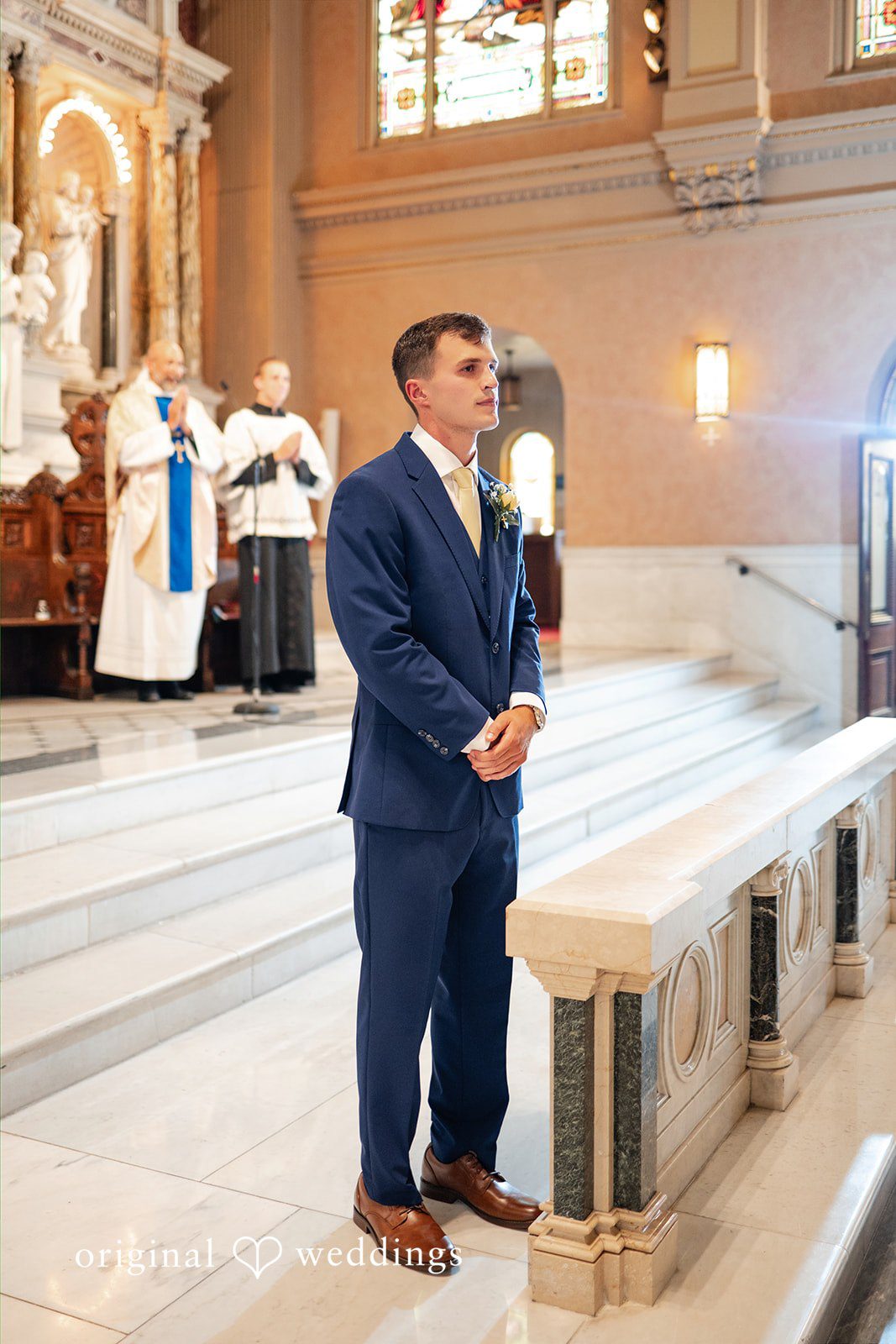 A groom in a blue suit stands near the wooden pews of a chapel aisle.