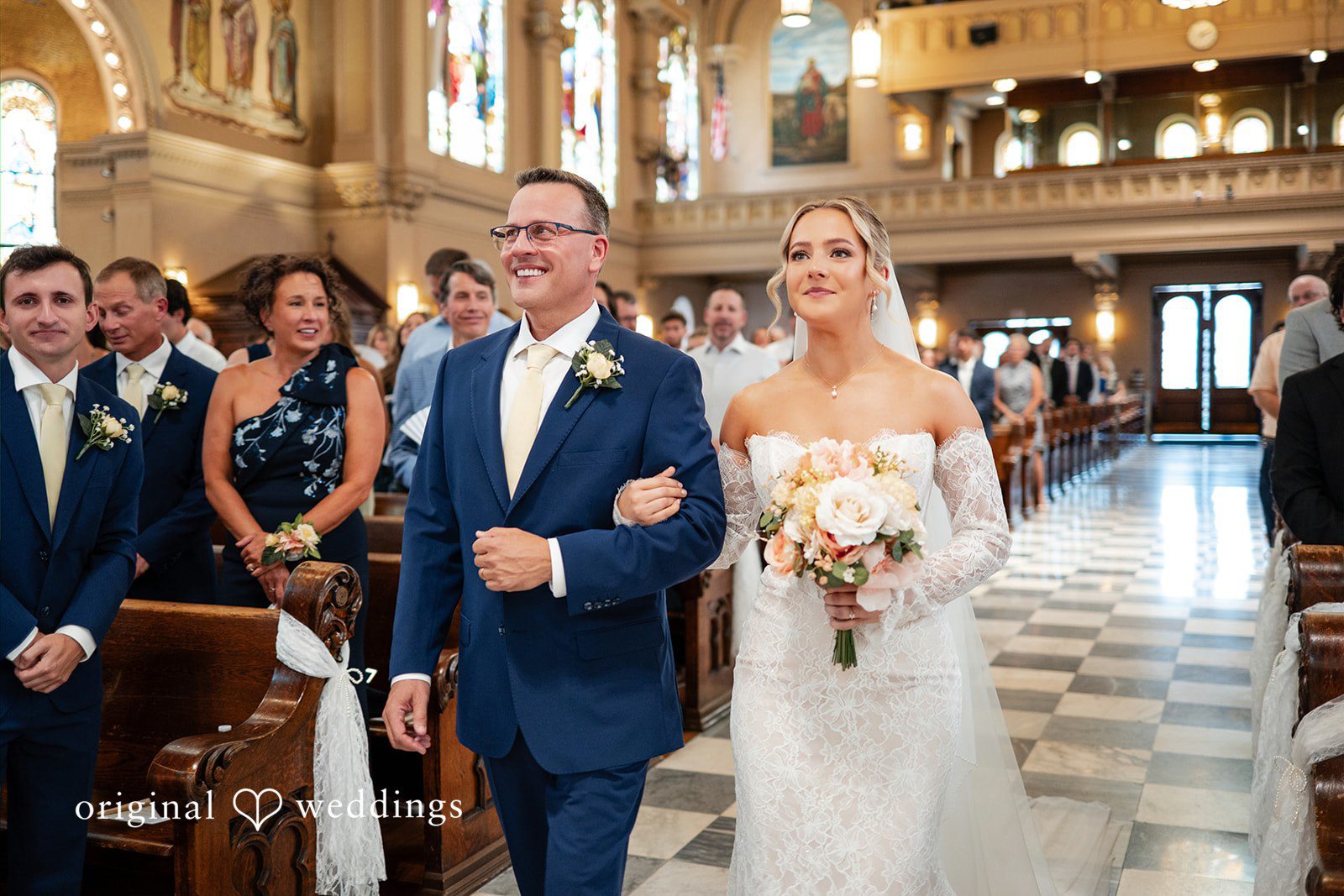 A bride in a lace wedding gown walks arm-in-arm down a church aisle with a man wearing glasses and a blue suit.