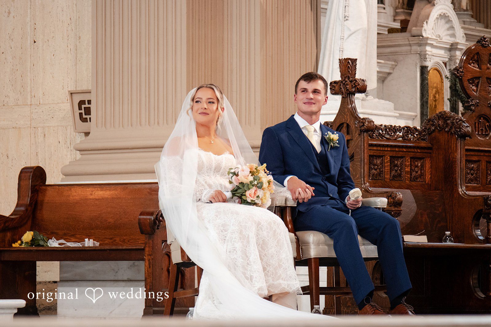 A bride sits next to a groom in a dark suit on a wooden church pew.