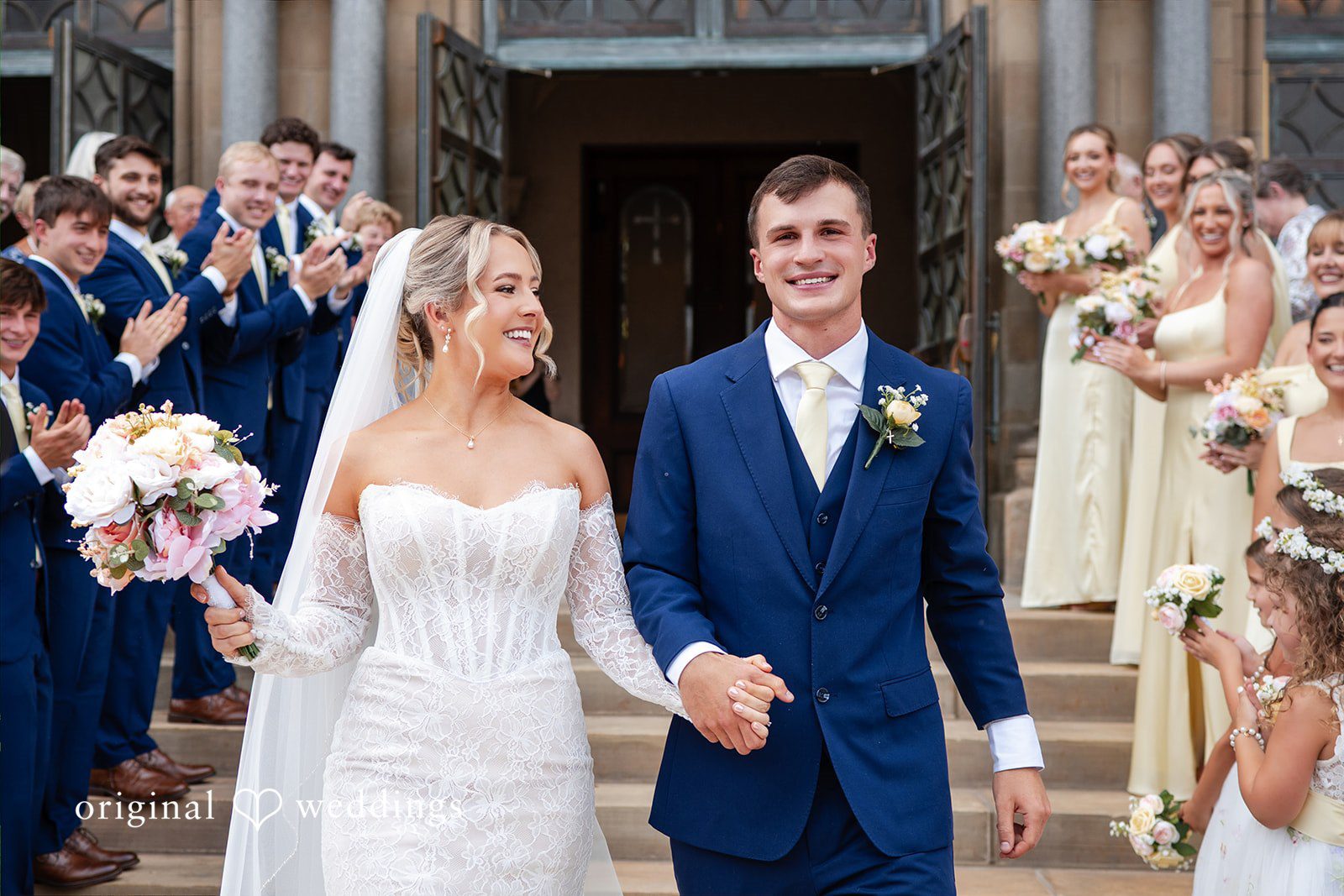 A bride and groom stand beside their wedding party against a stone background.