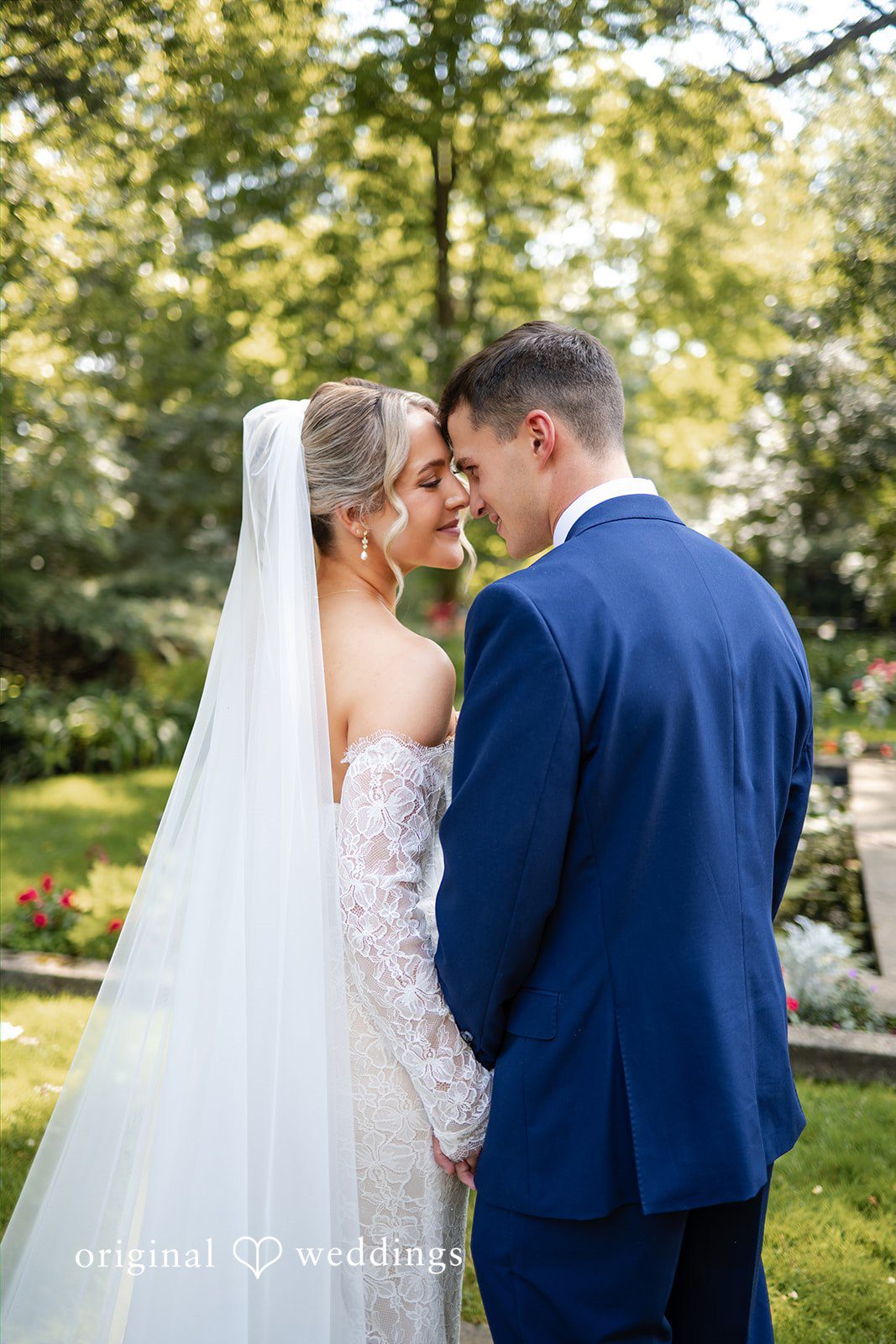 A bride in a long veil stands beside a groom in a blue suit with their backs to the camera.