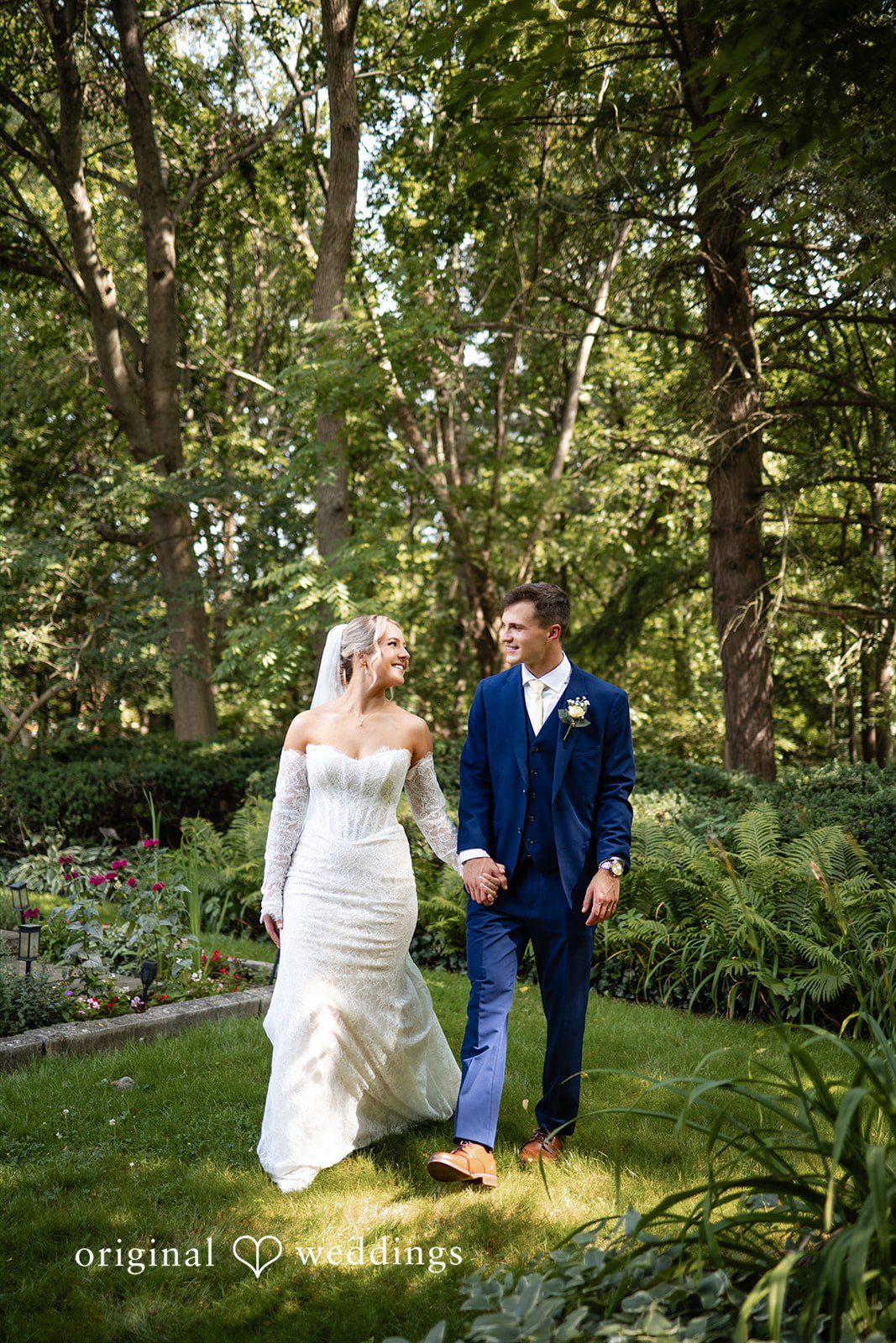 A bride and groom walk together along a path winding through a wooded garden area.