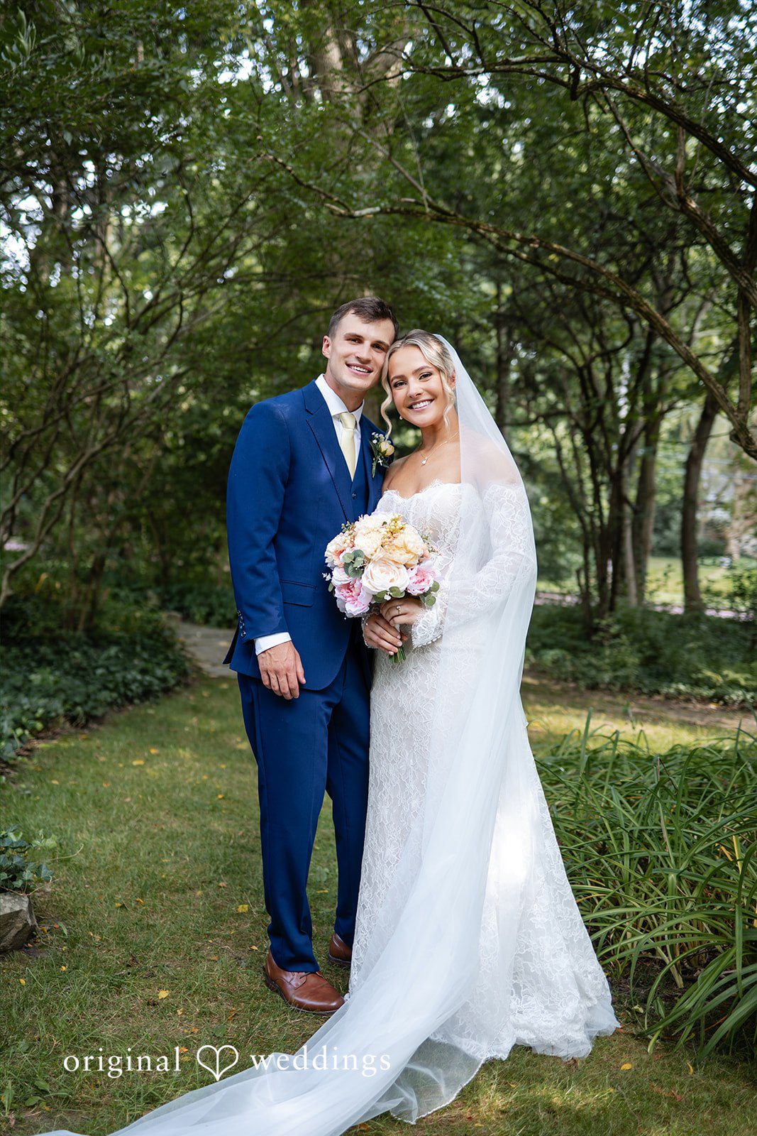 A bride and groom stand close together and smile on a grassy lawn surrounded by trees.