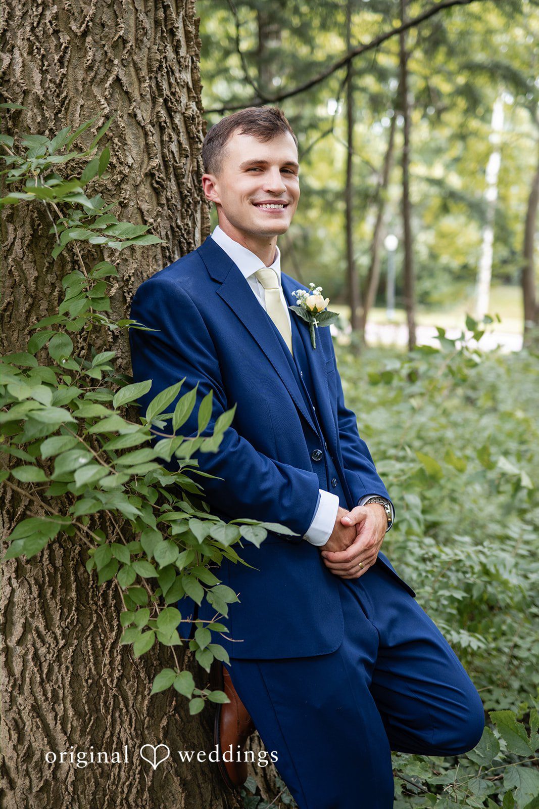 A man in a blue suit leans back while posing among lush green foliage.