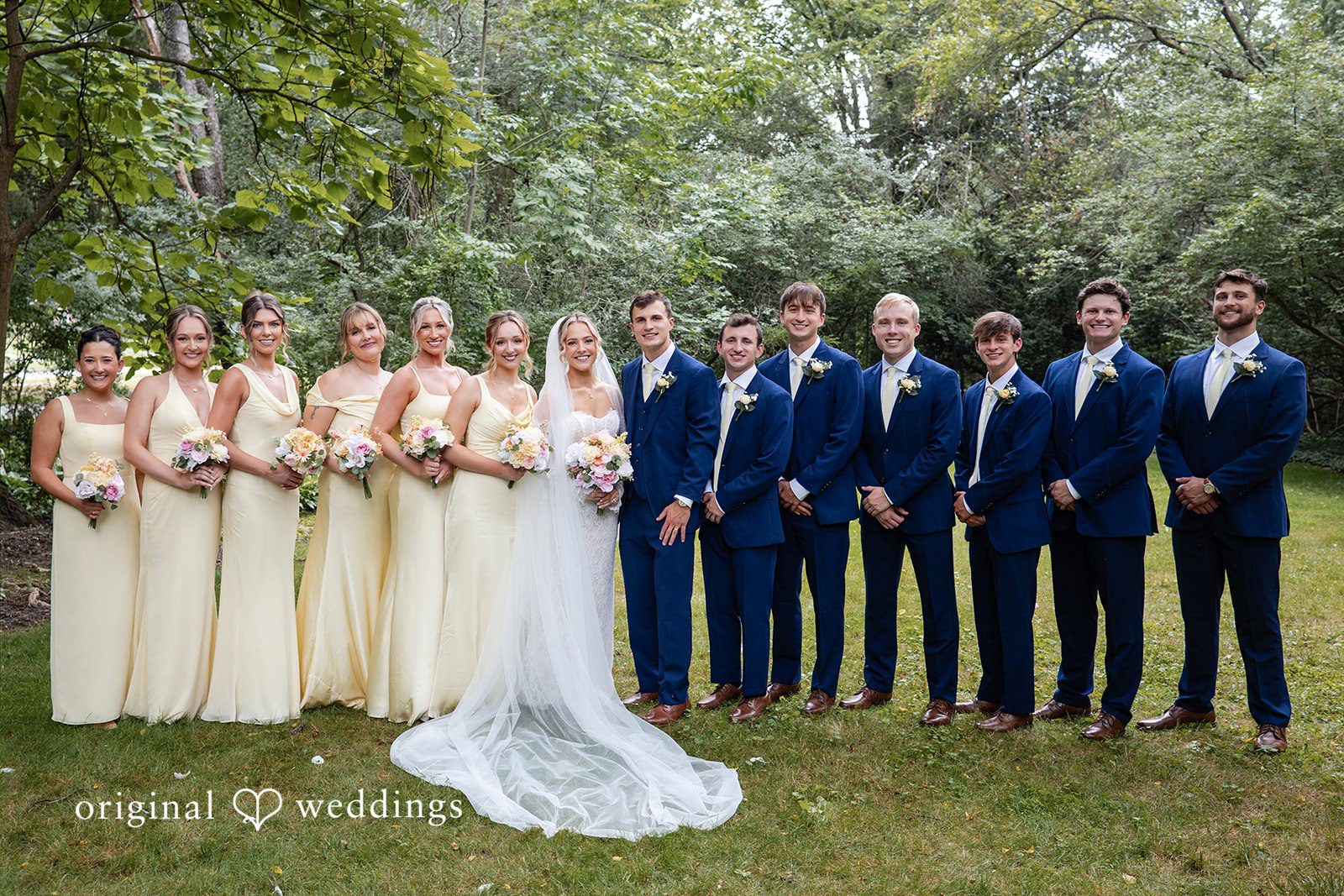A full wedding party poses together in a long line on a grassy field.