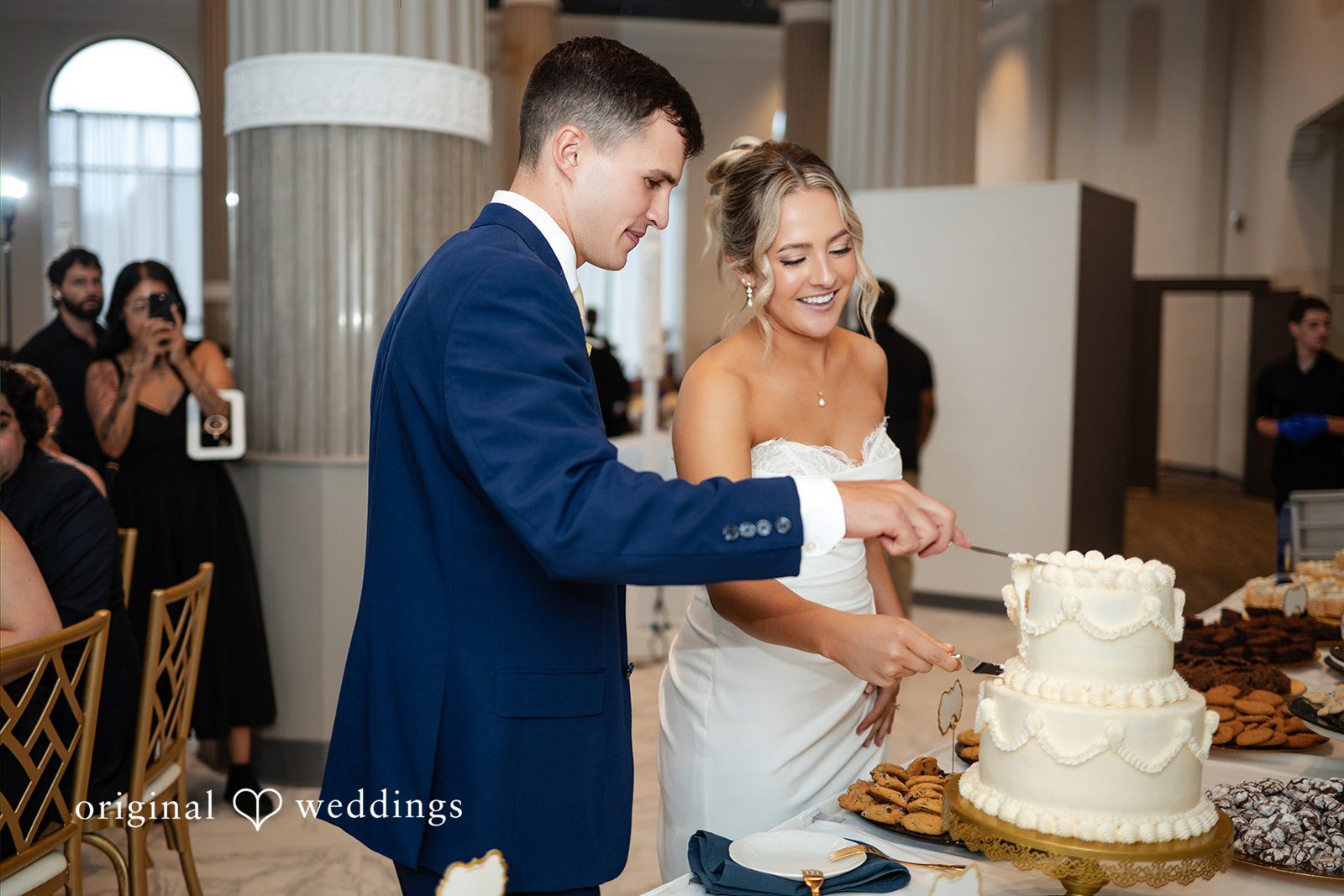 A bride and groom stand together to cut their multi-tiered white wedding cake.