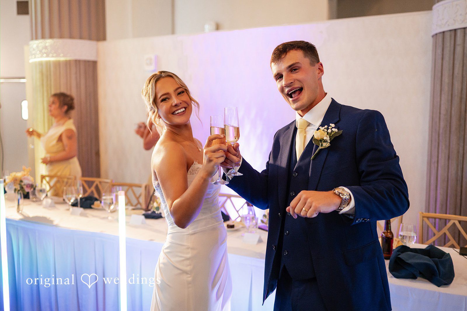 A bride and groom smile joyfully while dancing together against a brightly lit backdrop.
