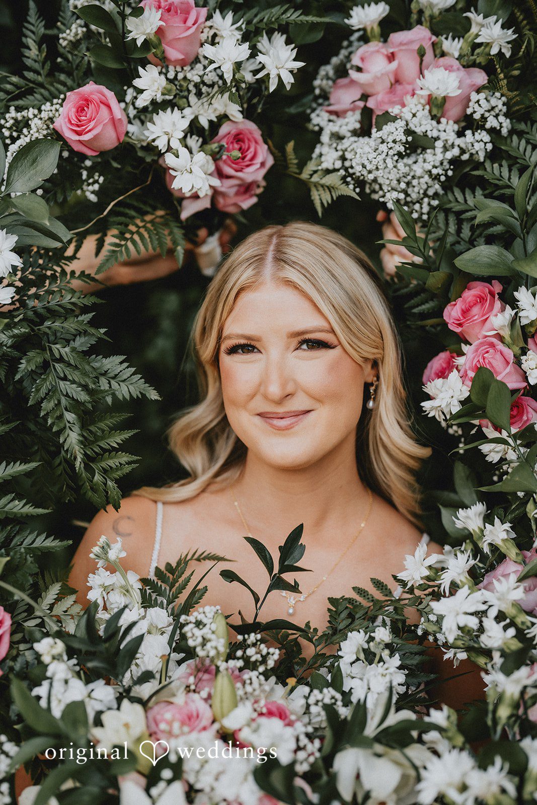 Ashley + Dylan A smiling blonde bride posing in front of a vibrant wall of pink and purple flowers.