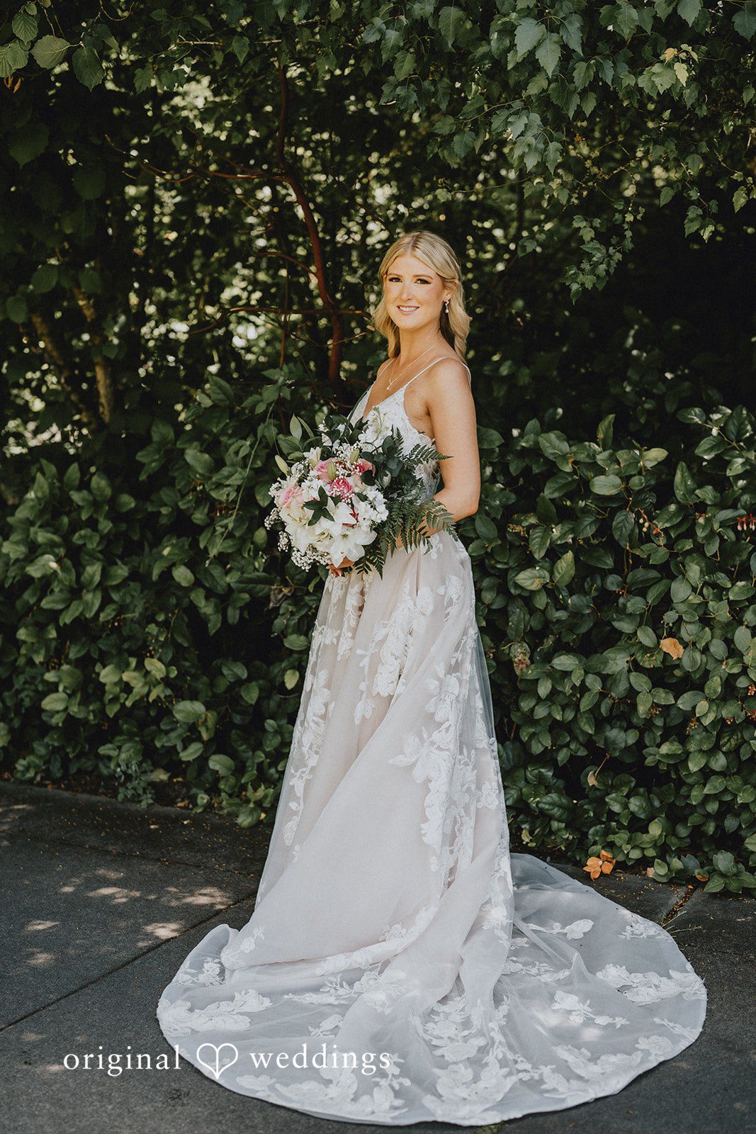 Ashley + Dylan A bride in a white lace wedding dress standing alone on a grassy field.