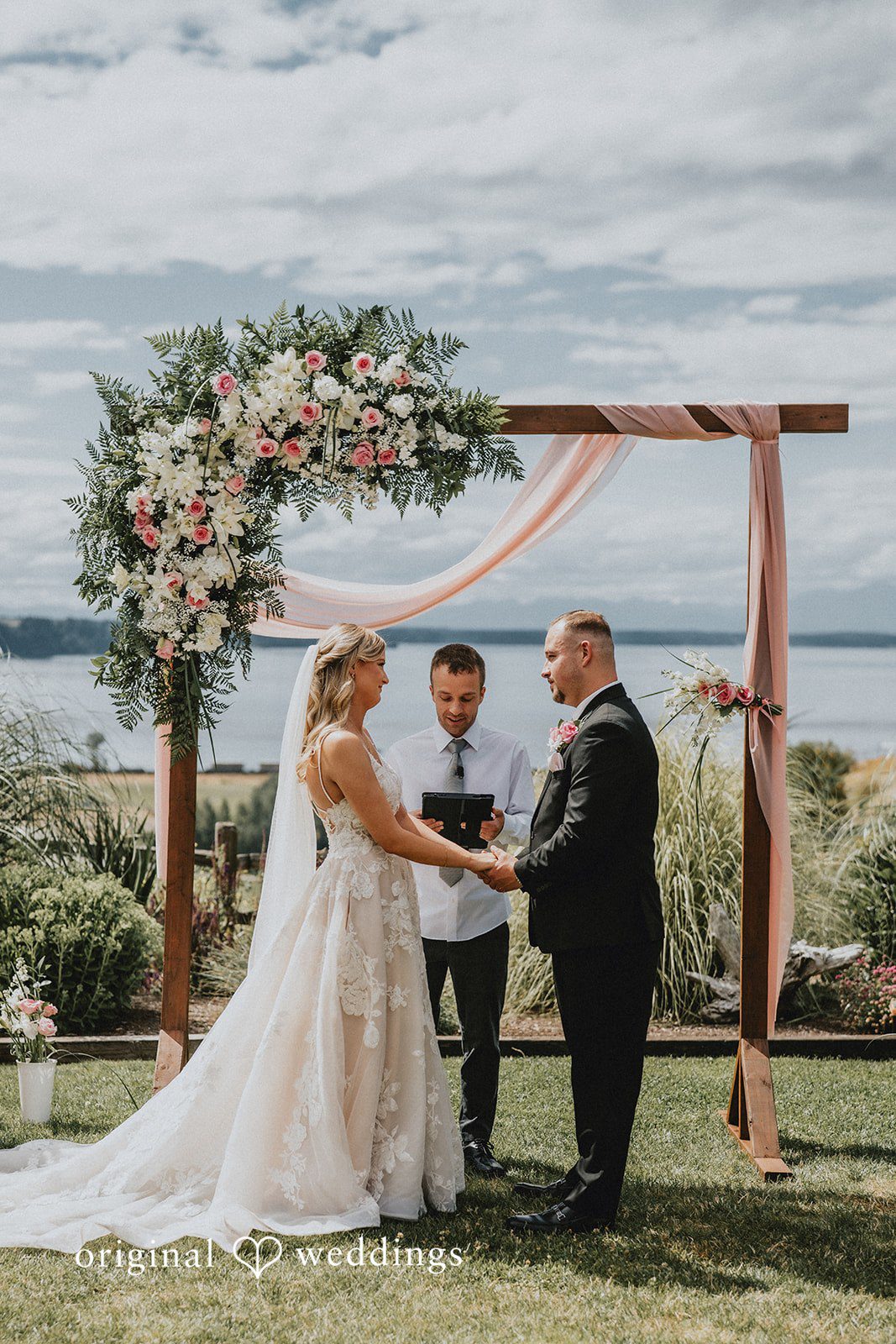 Ashley + Dylan A wedding ceremony in progress under a wooden arch decorated with white drapery and greenery.