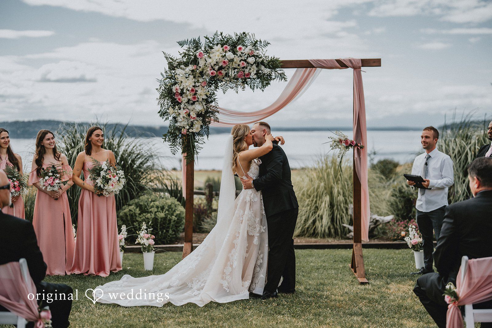Ashley + Dylan A wide view of a bride and groom standing under a rustic wooden arch next to a lake.