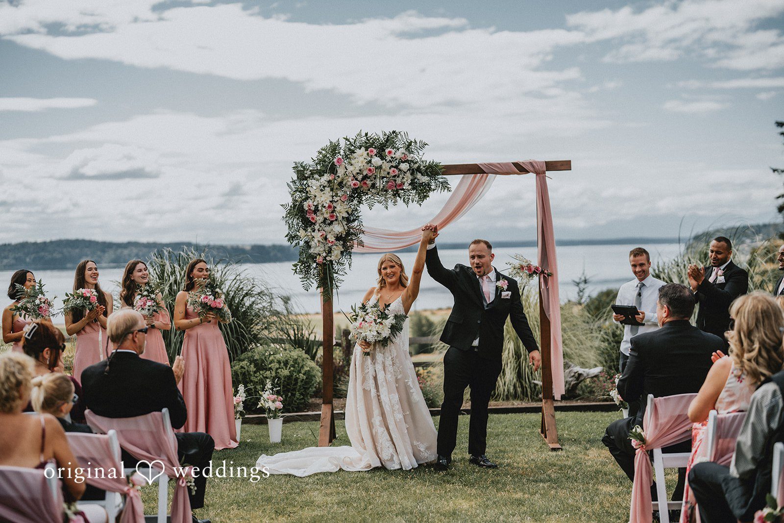 Ashley + Dylan A newly married bride and groom holding hands and raising their arms in celebration under a floral wooden arch.
