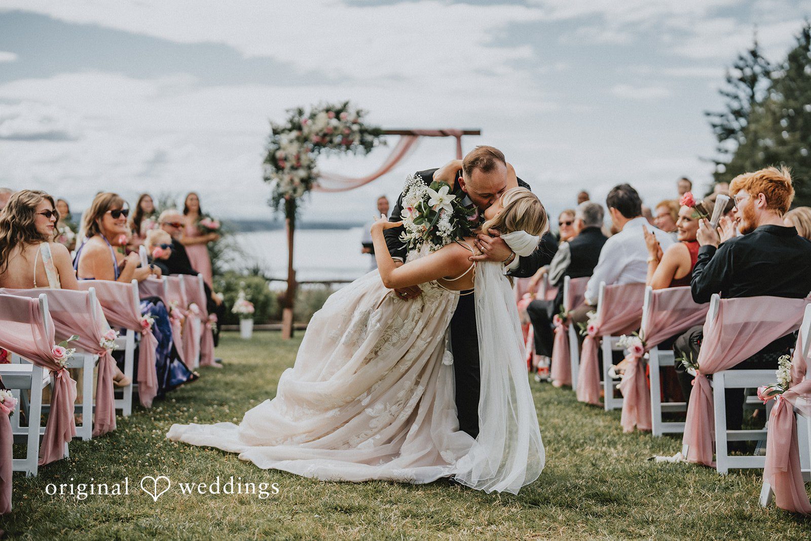 Ashley + Dylan A bride and groom sharing a kiss under a floral arch surrounded by wedding guests.