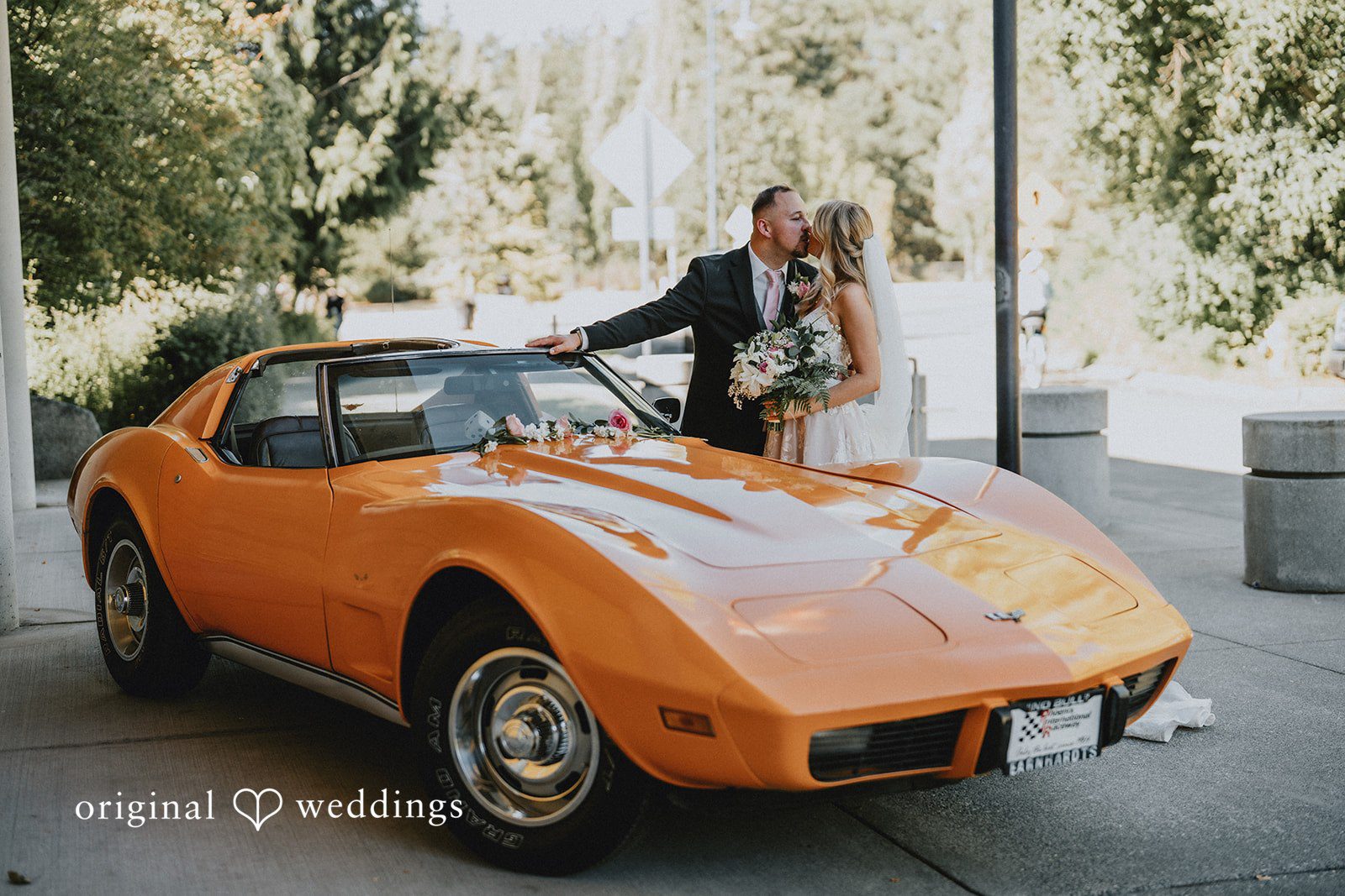 Ashley + Dylan An orange vintage sports car parked on a paved driveway with a couple nearby.