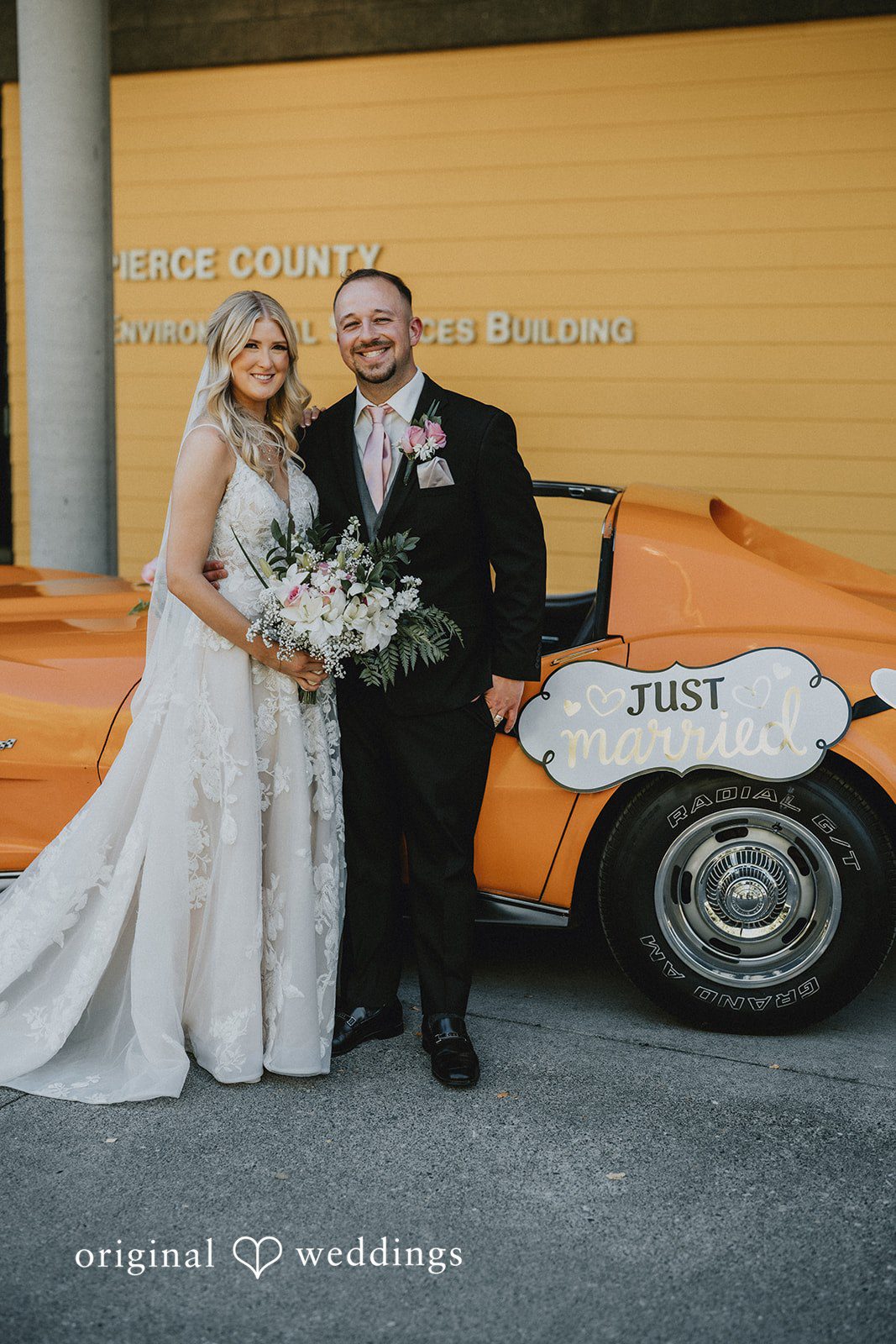 Ashley + Dylan A bride and groom posing together in front of a bright yellow wall next to a vintage car.