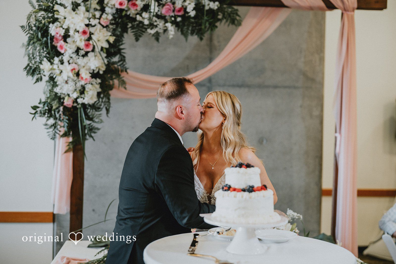Ashley + Dylan A bride and groom sharing a romantic kiss behind a small two-tiered wedding cake decorated with fresh berries.